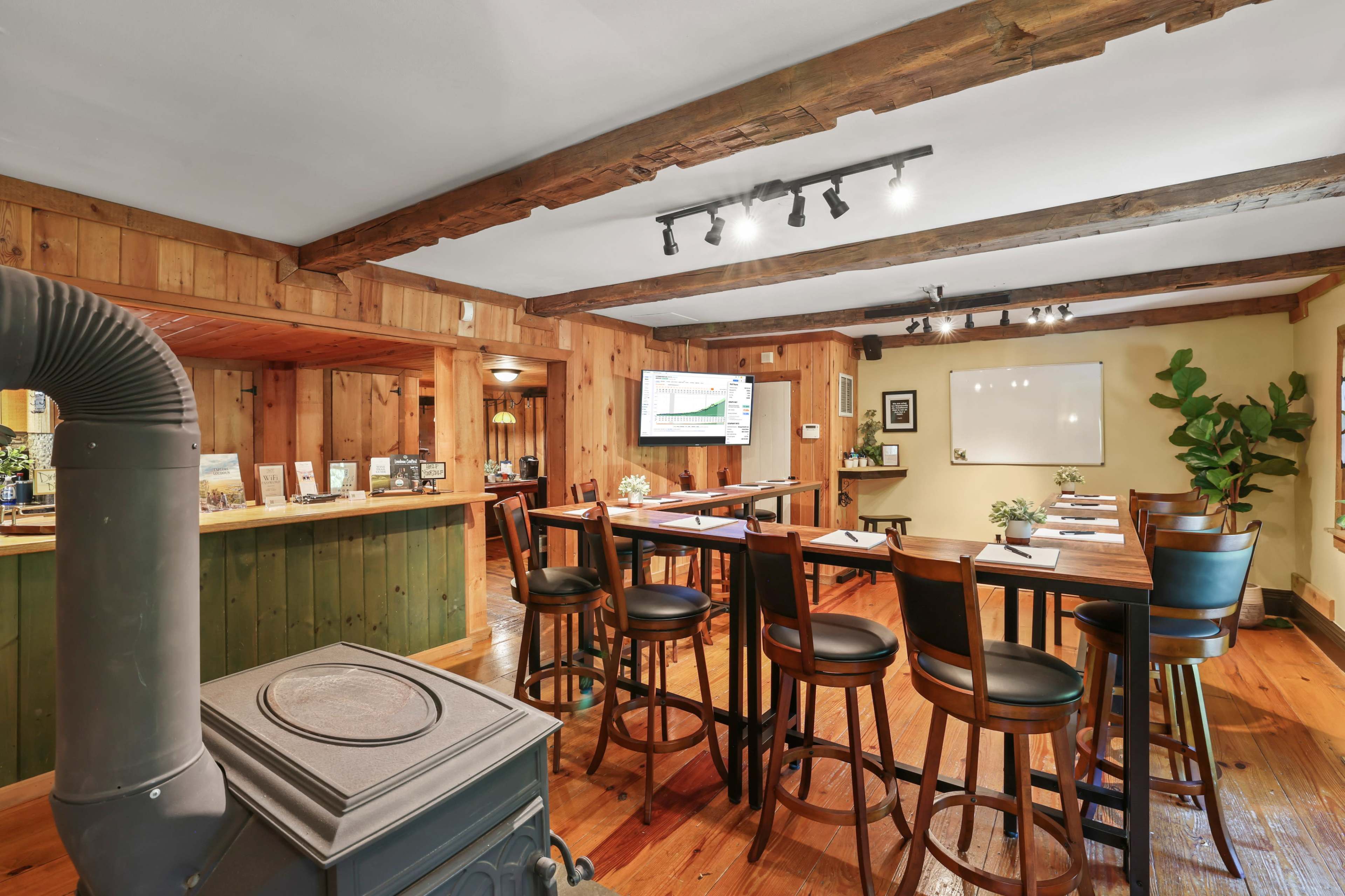 A rustic meeting room featuring wooden beams, a bar area, several tables set up for a presentation, and a large screen on the wall.