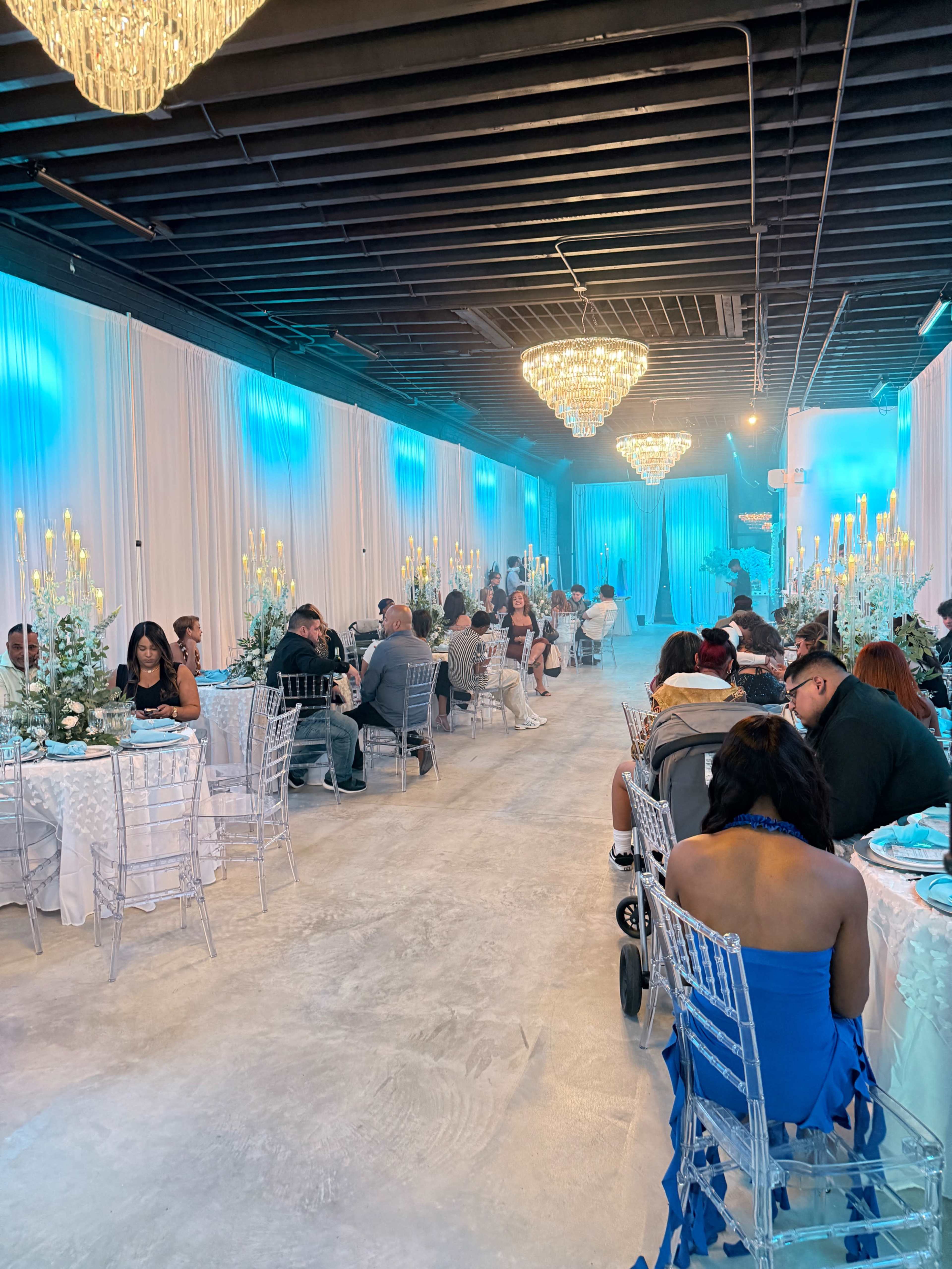 The image shows a spacious event hall decorated with teal lighting and chandeliers, where guests are seated at elegantly set tables for a gathering or celebration.