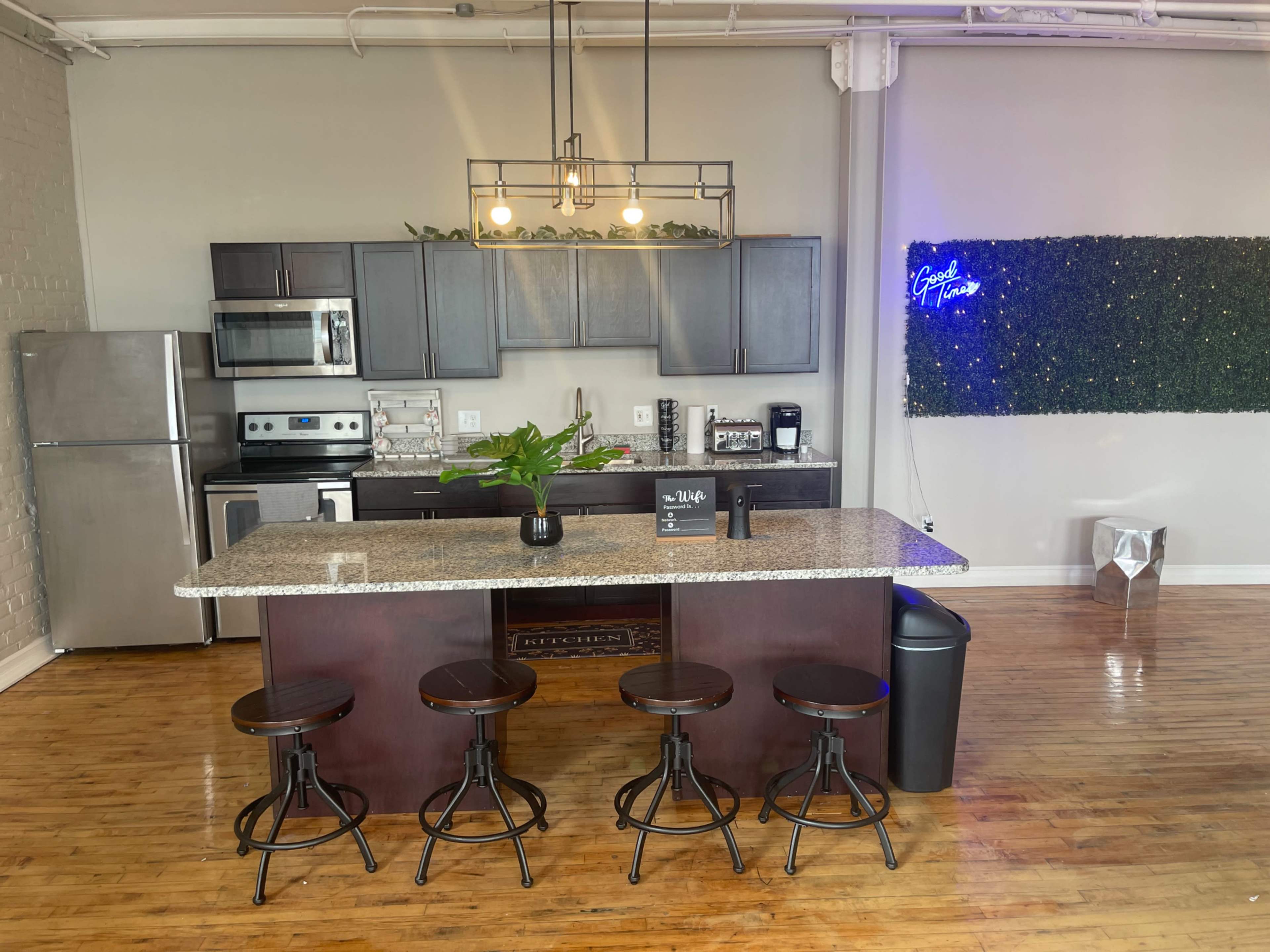 The image shows a modern kitchen with a granite island, stainless steel appliances, and four tall stools, set against a backdrop of a decorative green wall.