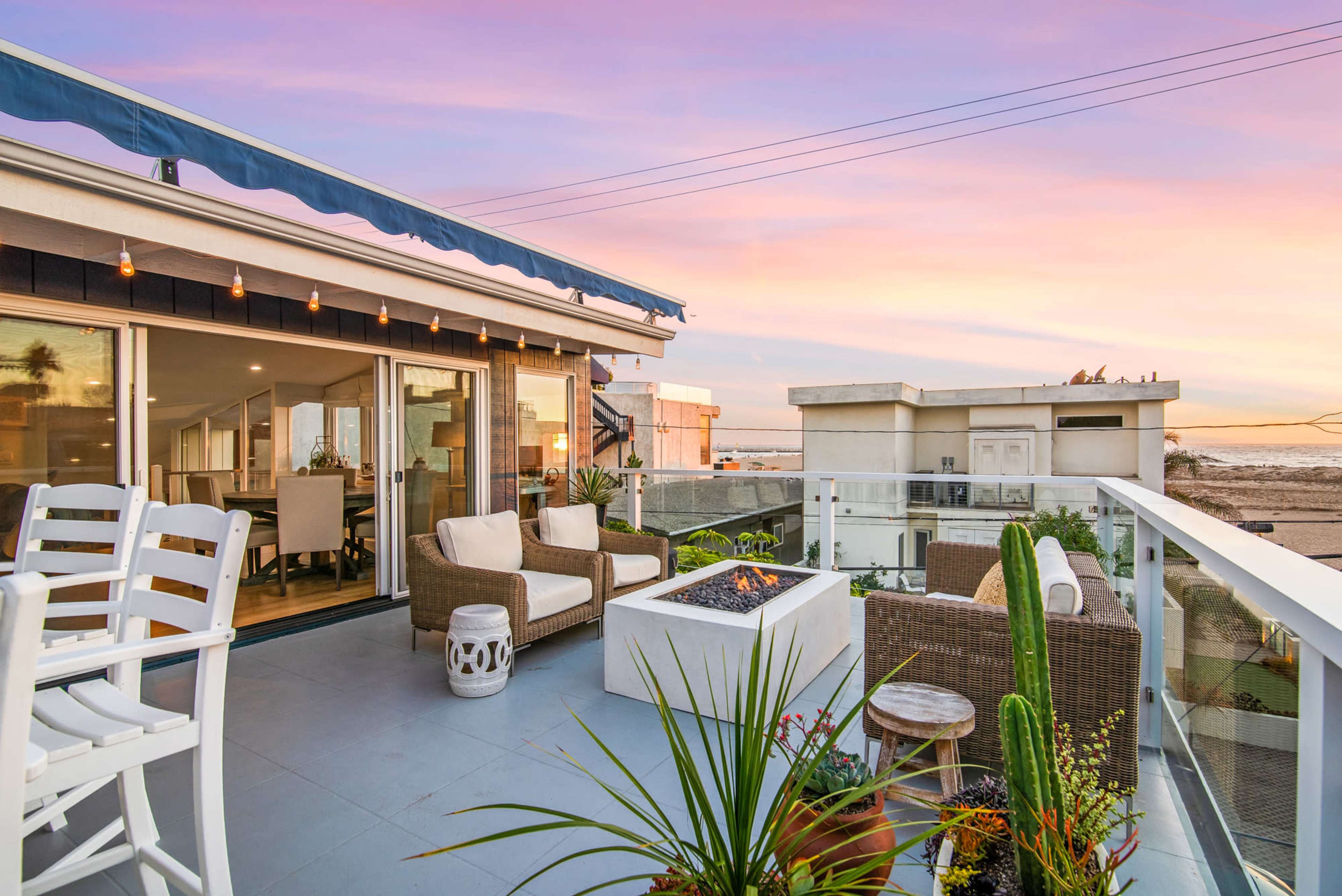 A modern patio overlooks a beach at sunset, featuring outdoor seating and a fire pit surrounded by potted plants.
