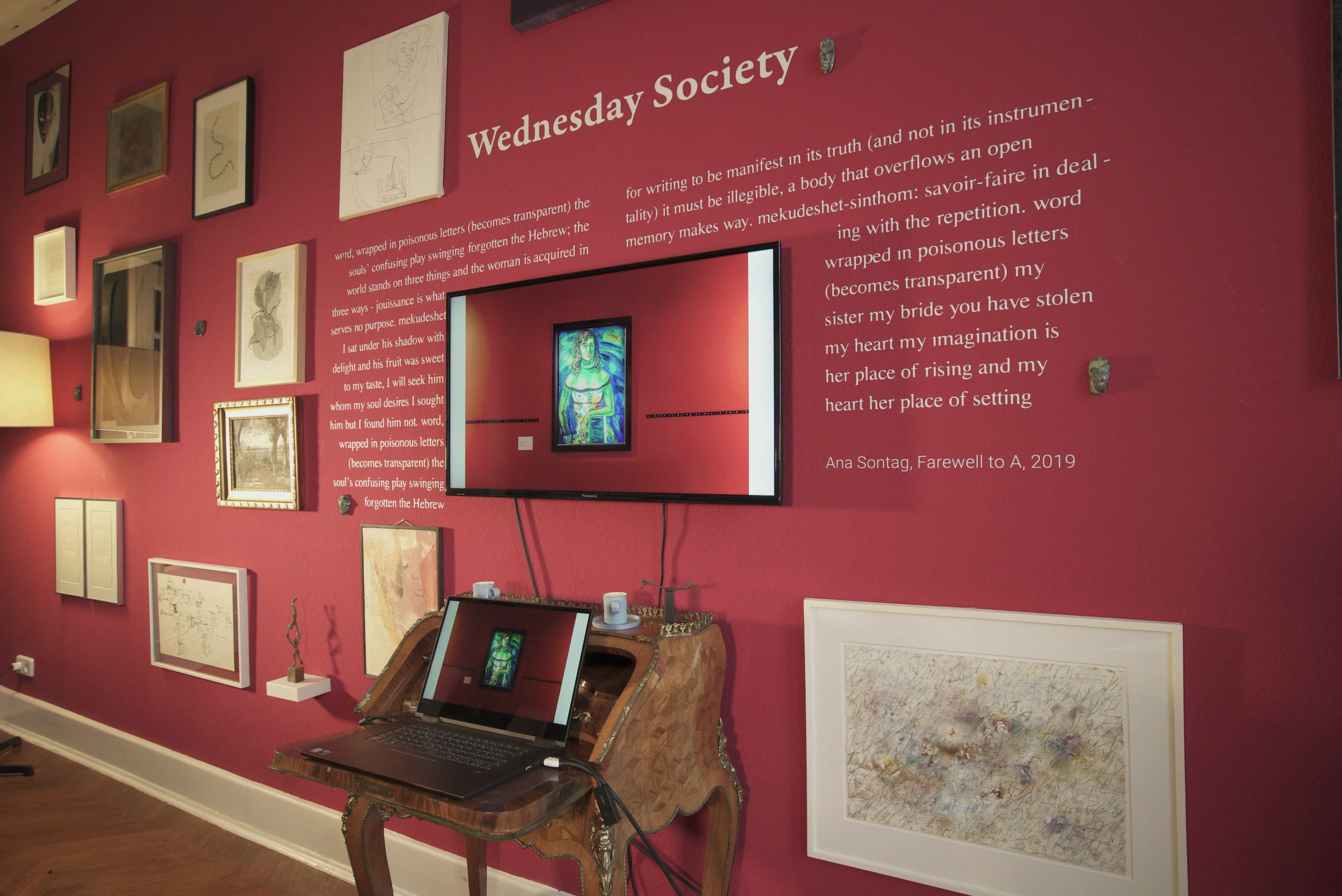 The image showcases a room with a rich maroon wall adorned with various framed artworks and a central television displaying a piece, accompanied by a desk with a laptop.