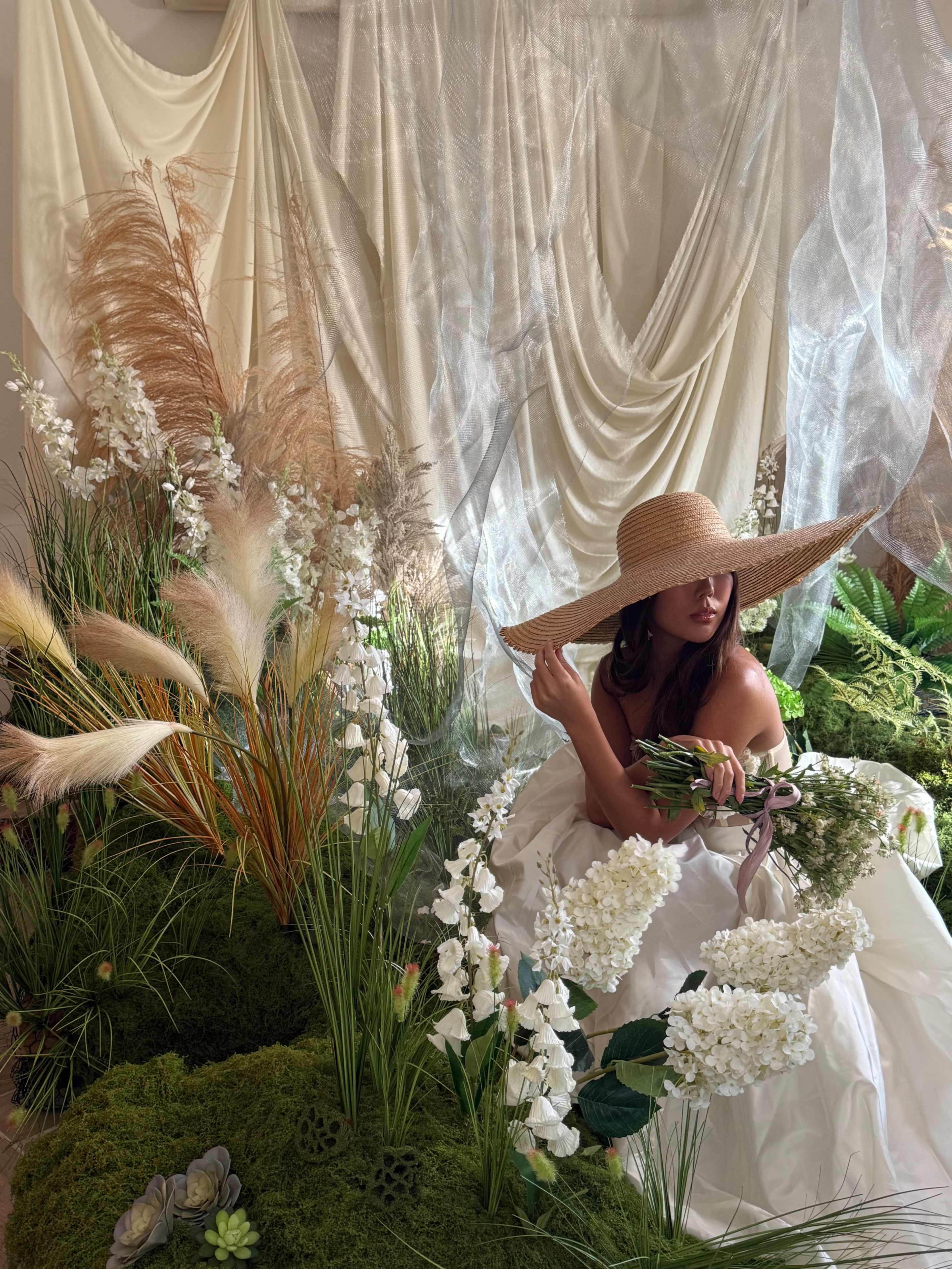 A woman in a wide-brimmed straw hat poses among a lush arrangement of flowers and greenery.