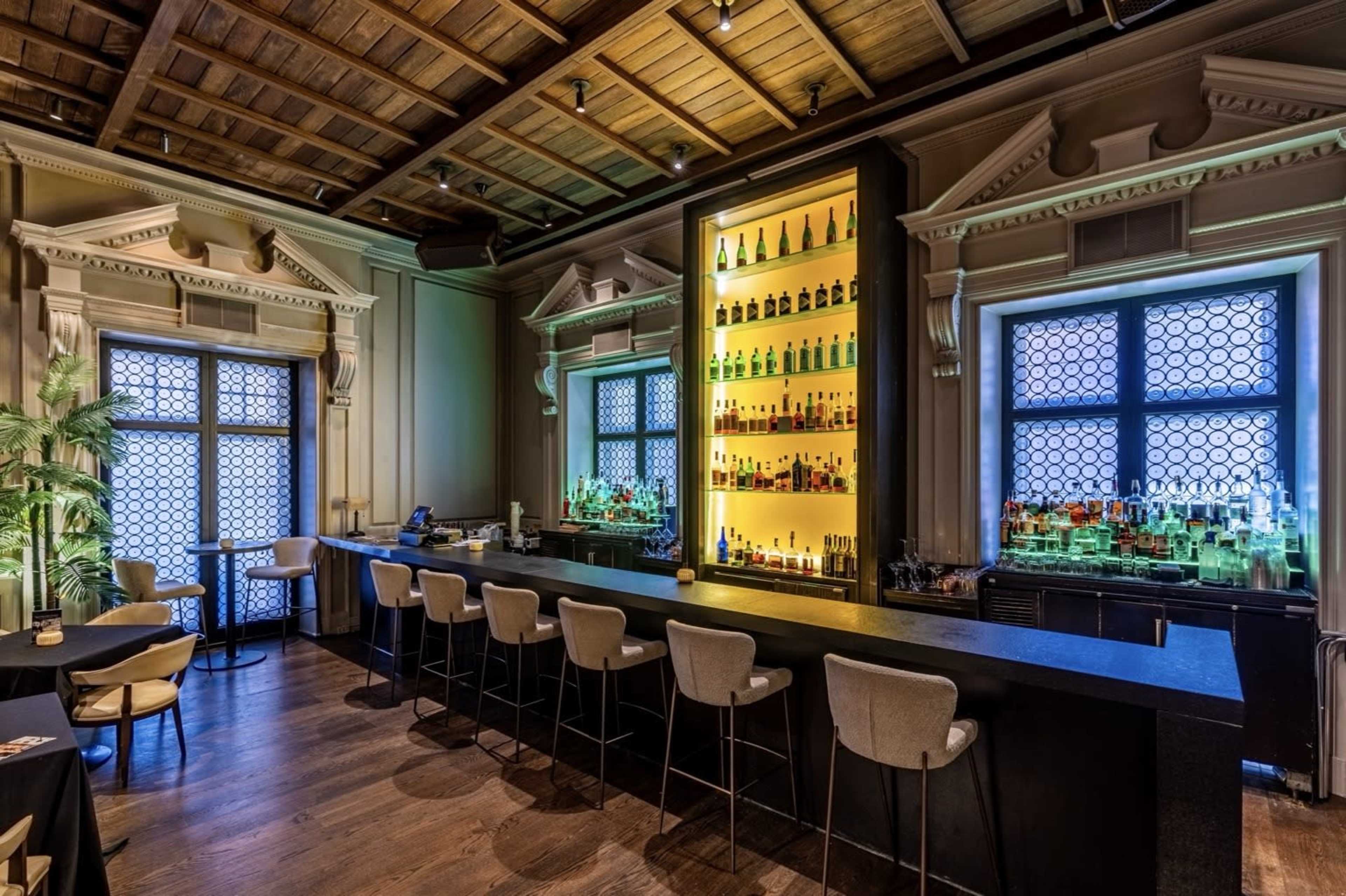 The image shows a stylish bar area with a backlit shelf of bottles, surrounded by elegant architectural details and seating at a countertop.