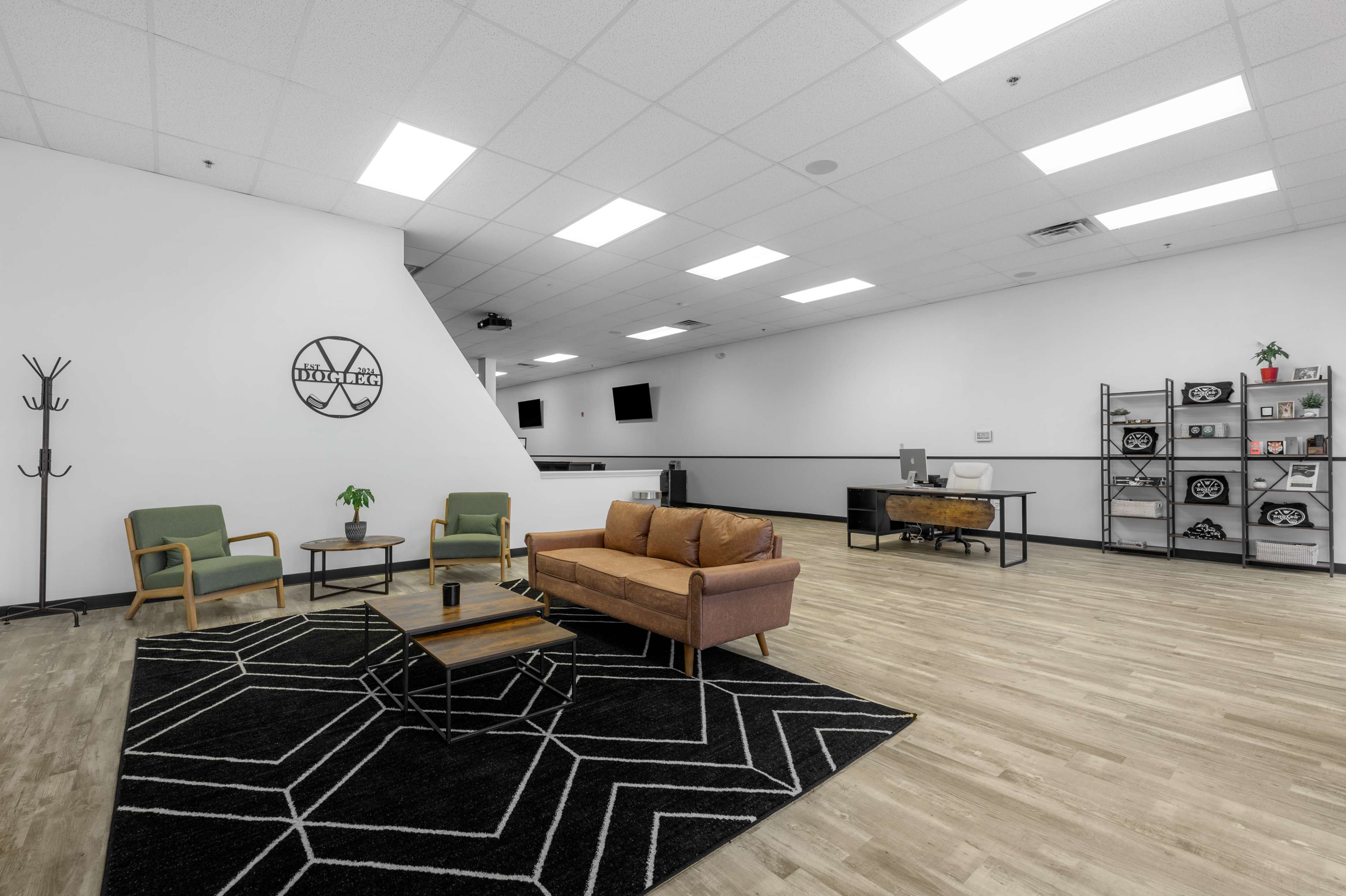 The image shows a spacious, modern office area featuring a brown sofa, two green chairs, a coffee table on a patterned rug, and a desk with shelves in the background.