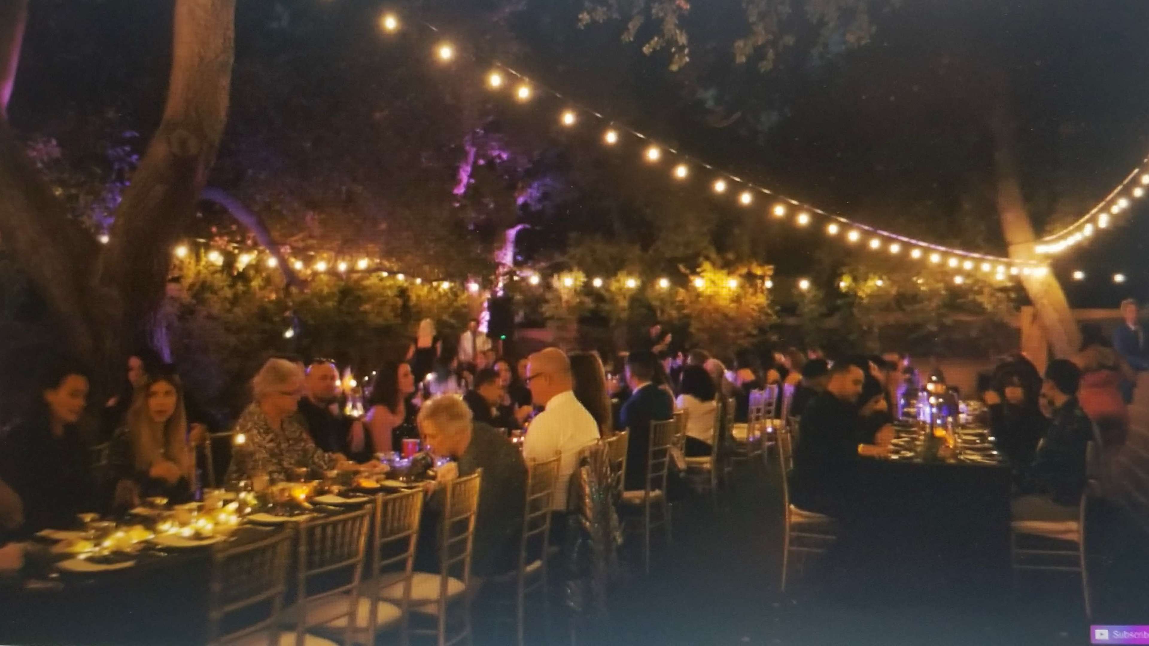 A long table is set for a dinner party outdoors, illuminated by string lights and surrounded by trees with guests seated on either side.