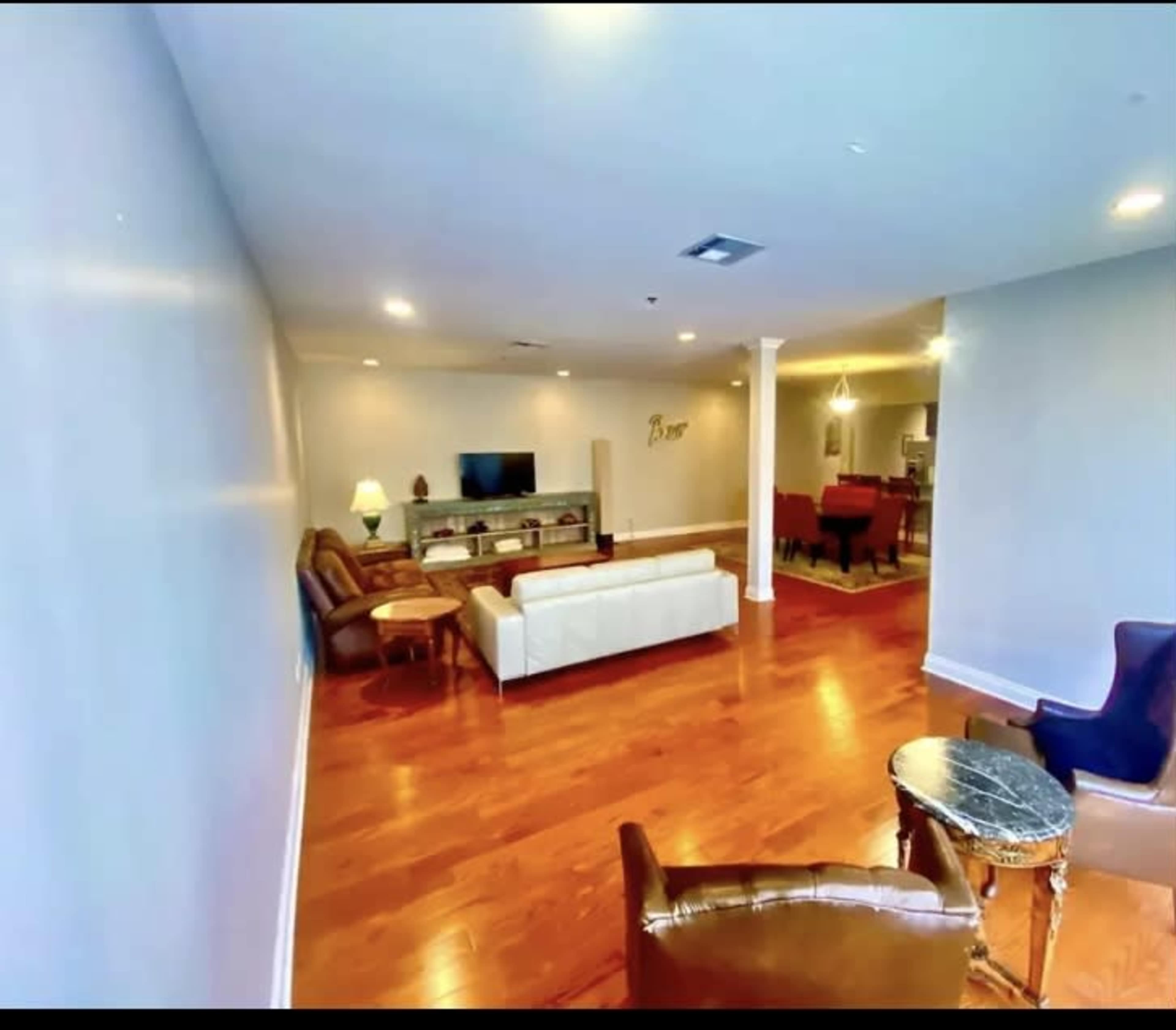 The image shows a spacious living area with wooden floors, a white sofa, two brown armchairs, and a television on a media console.