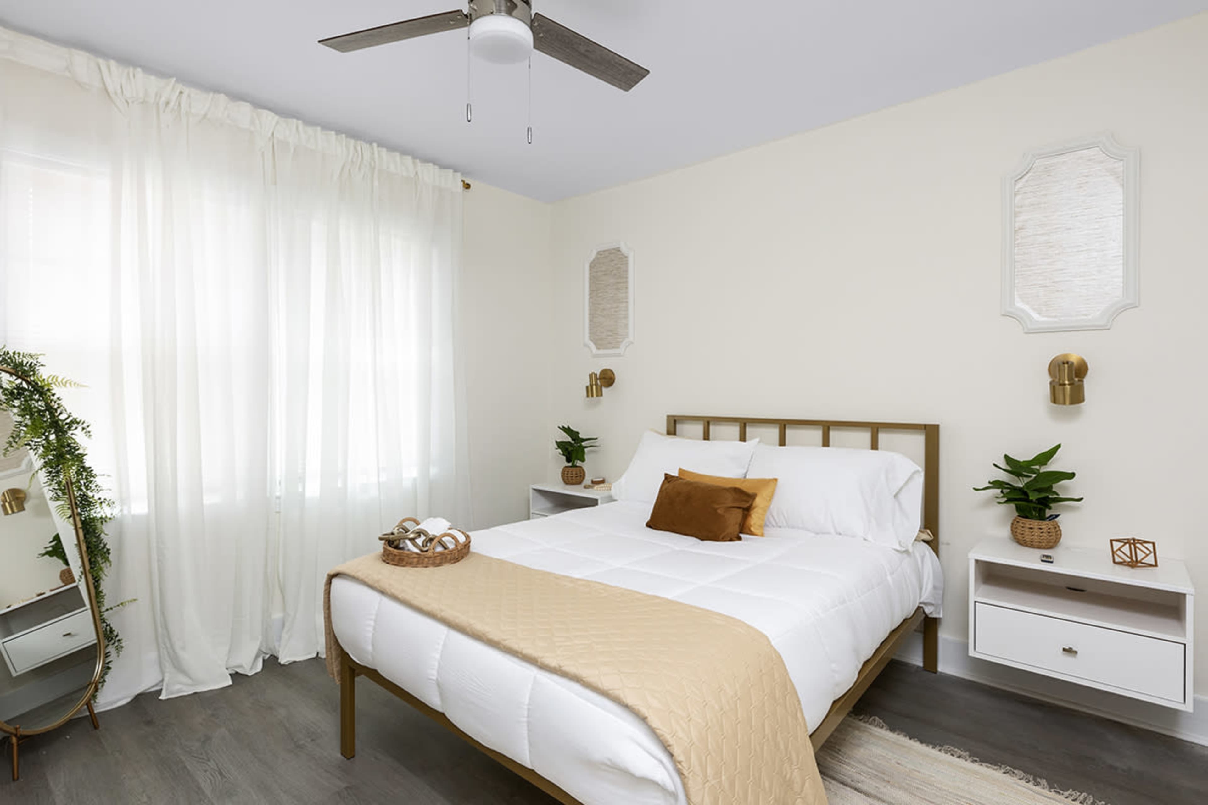 A bright bedroom features a double bed with a light-colored quilt, a nightstand on either side, and decorative plants on a dresser beneath sheer curtains.