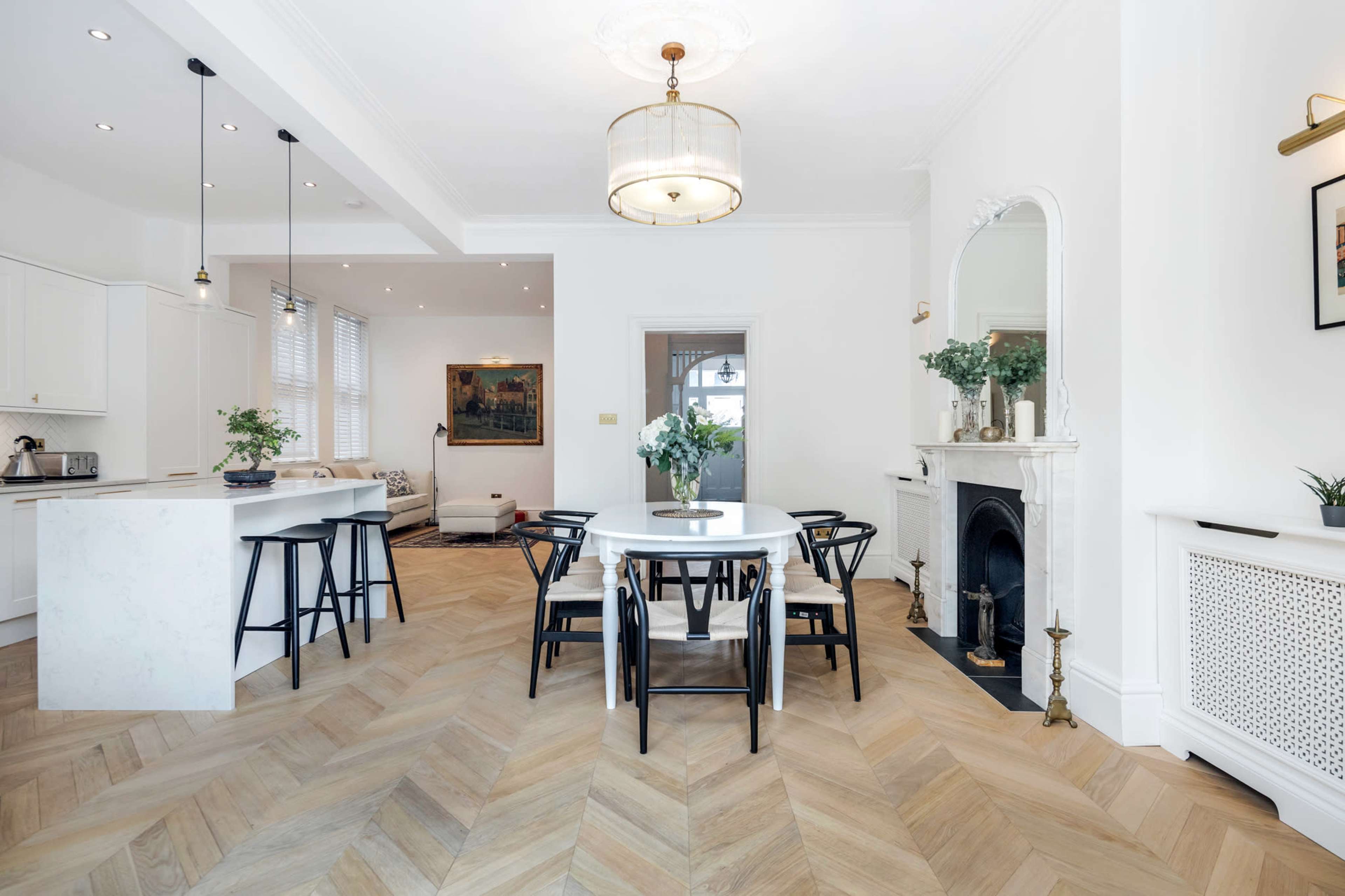 A modern kitchen and dining area features a round table with black chairs, a marble-topped island, and herringbone wood flooring beneath a stylish chandelier.