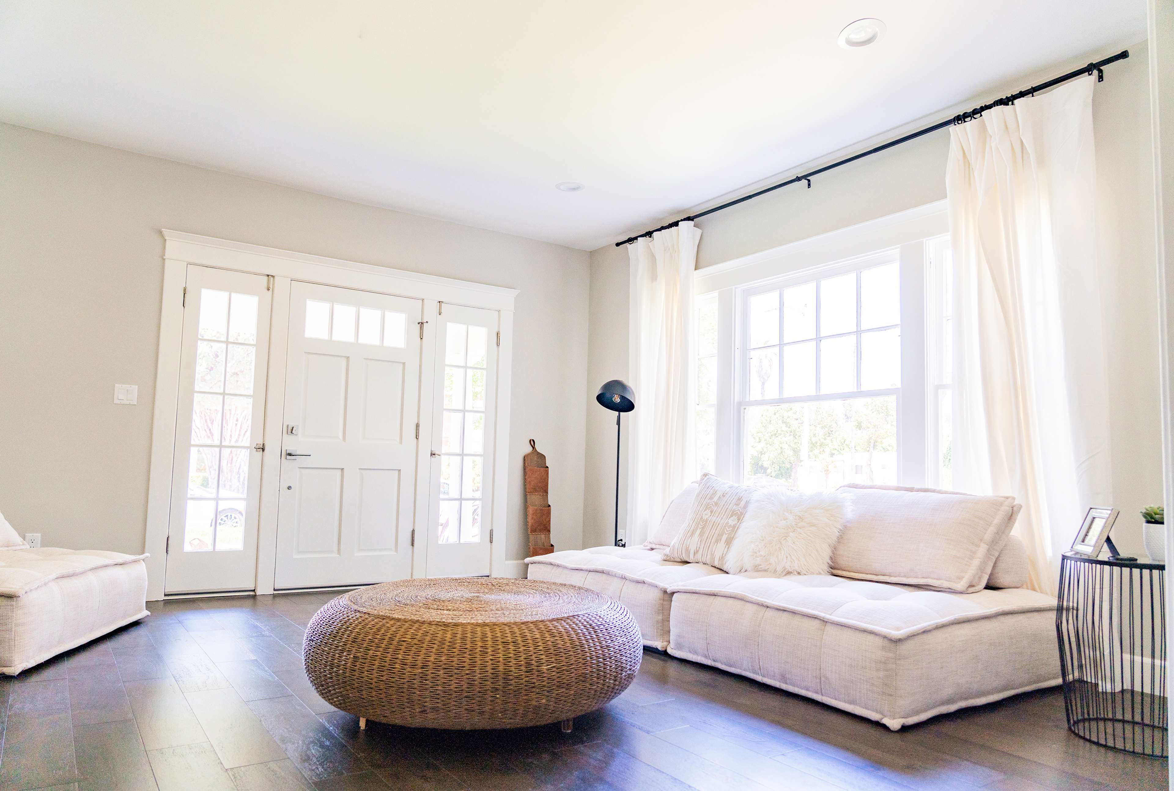 A bright living room with large windows, a round wicker coffee table, and a white sectional sofa against light gray walls.