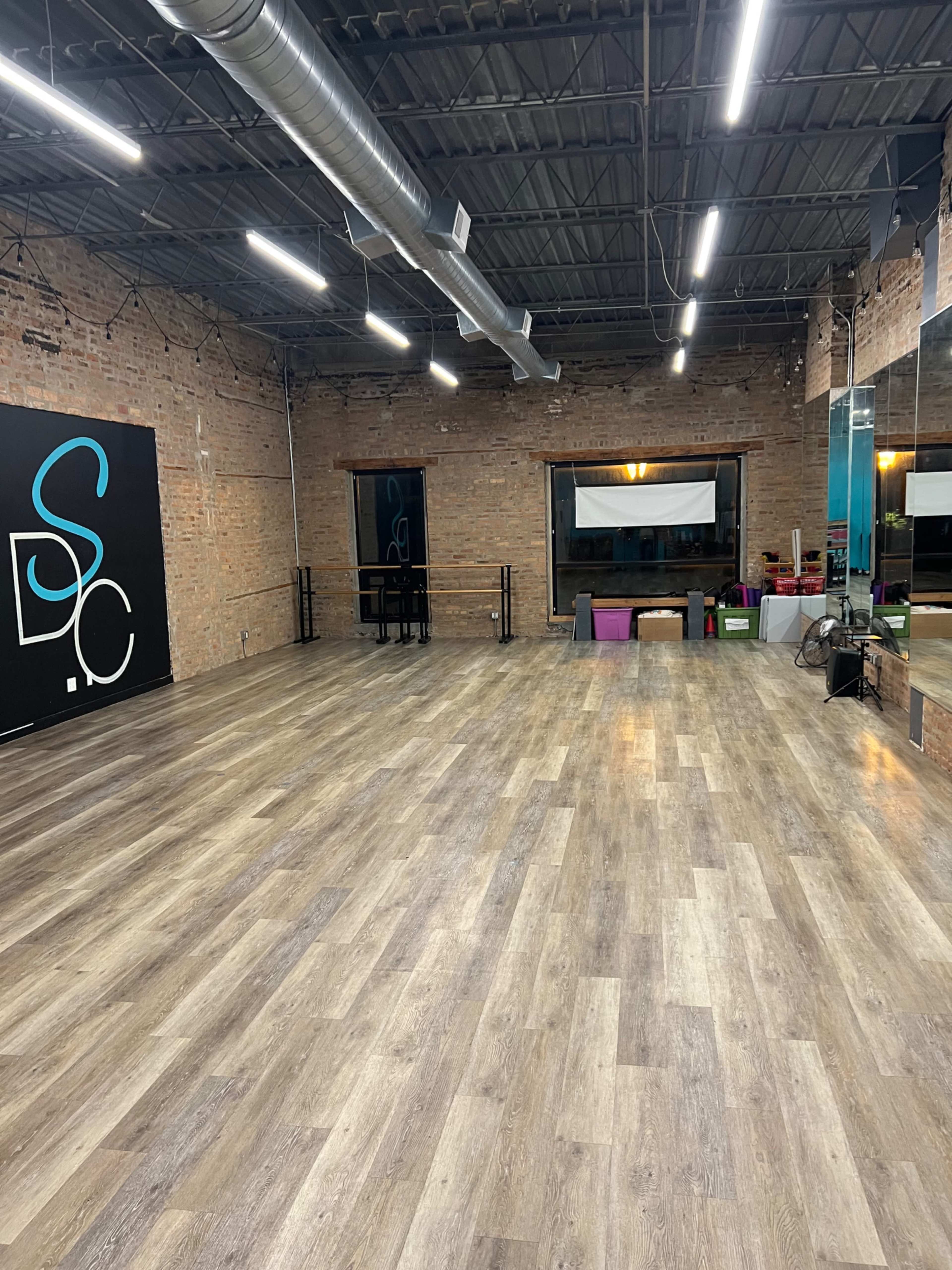 An empty dance studio with wooden flooring, exposed brick walls, and a large mirror reflecting the room's interior.