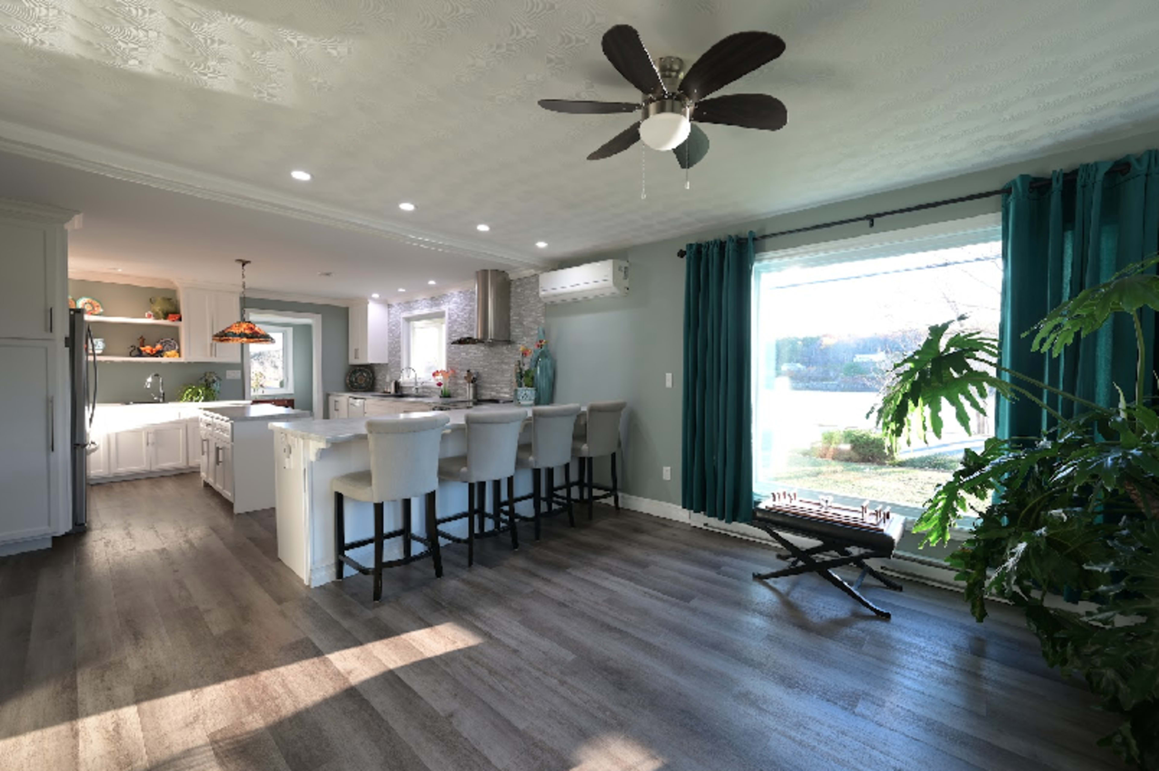 A modern kitchen with an island, bar stools, and a large window featuring teal curtains and a view of greenery outside.