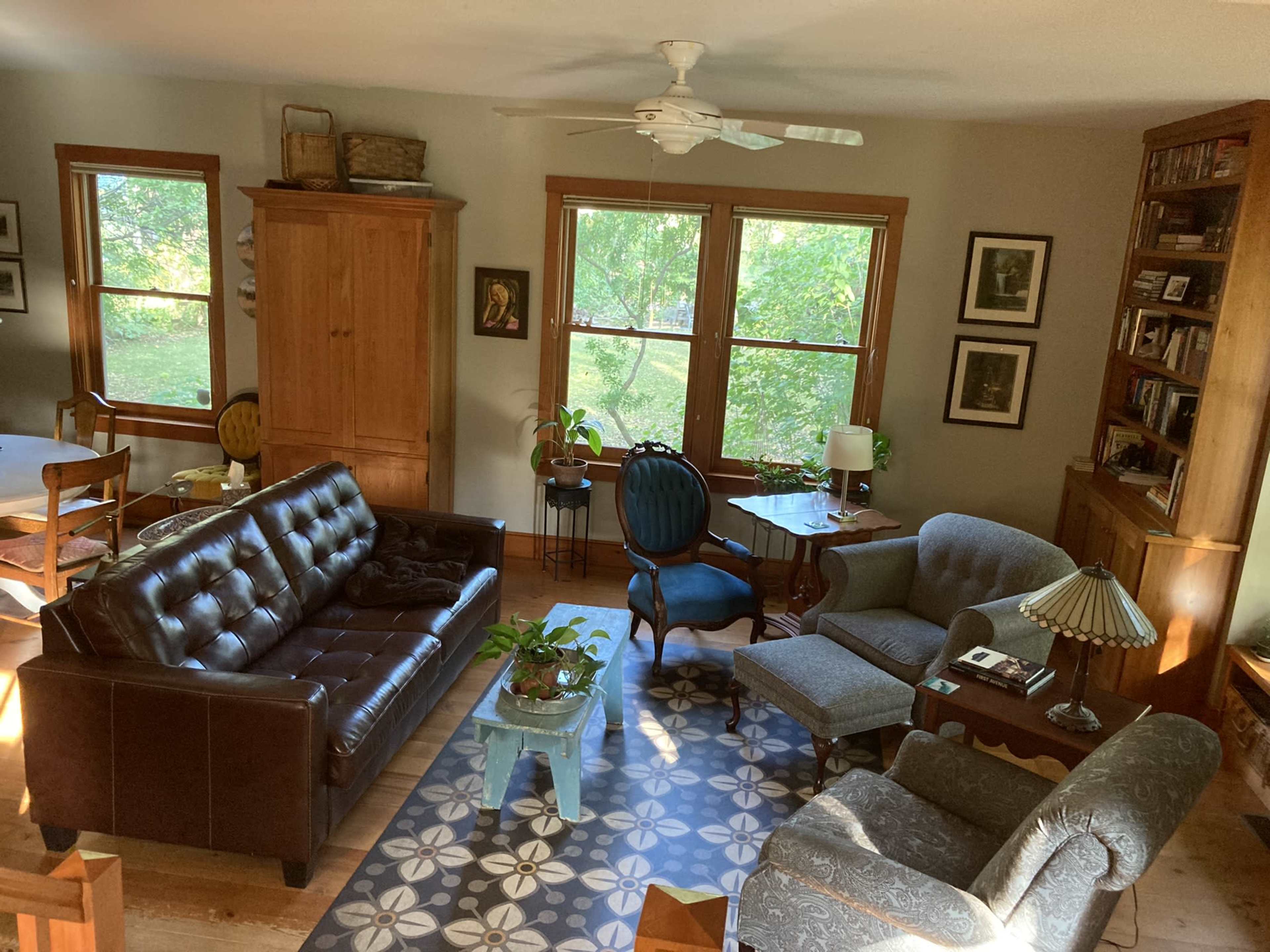 A cozy living room with a brown leather sofa, a blue armchair, and a wooden bookshelf, illuminated by natural light from large windows.