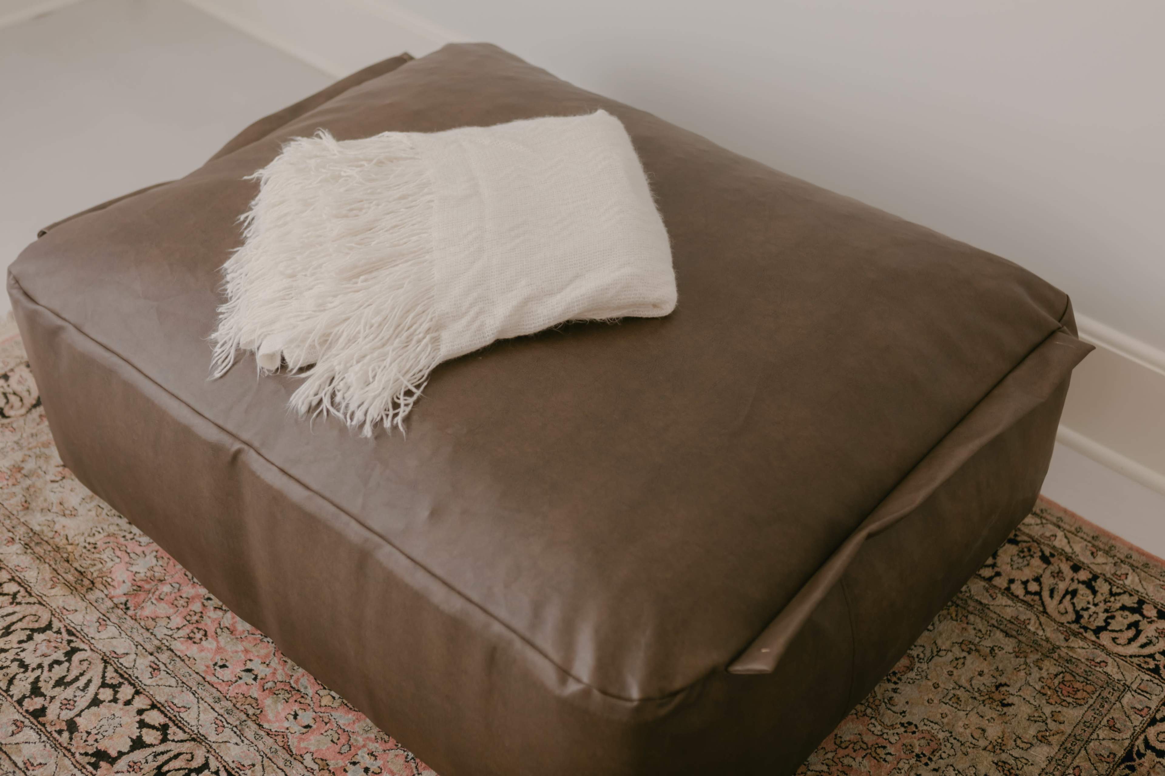 A large brown pouf is topped with a white fringed blanket, resting on a patterned rug.
