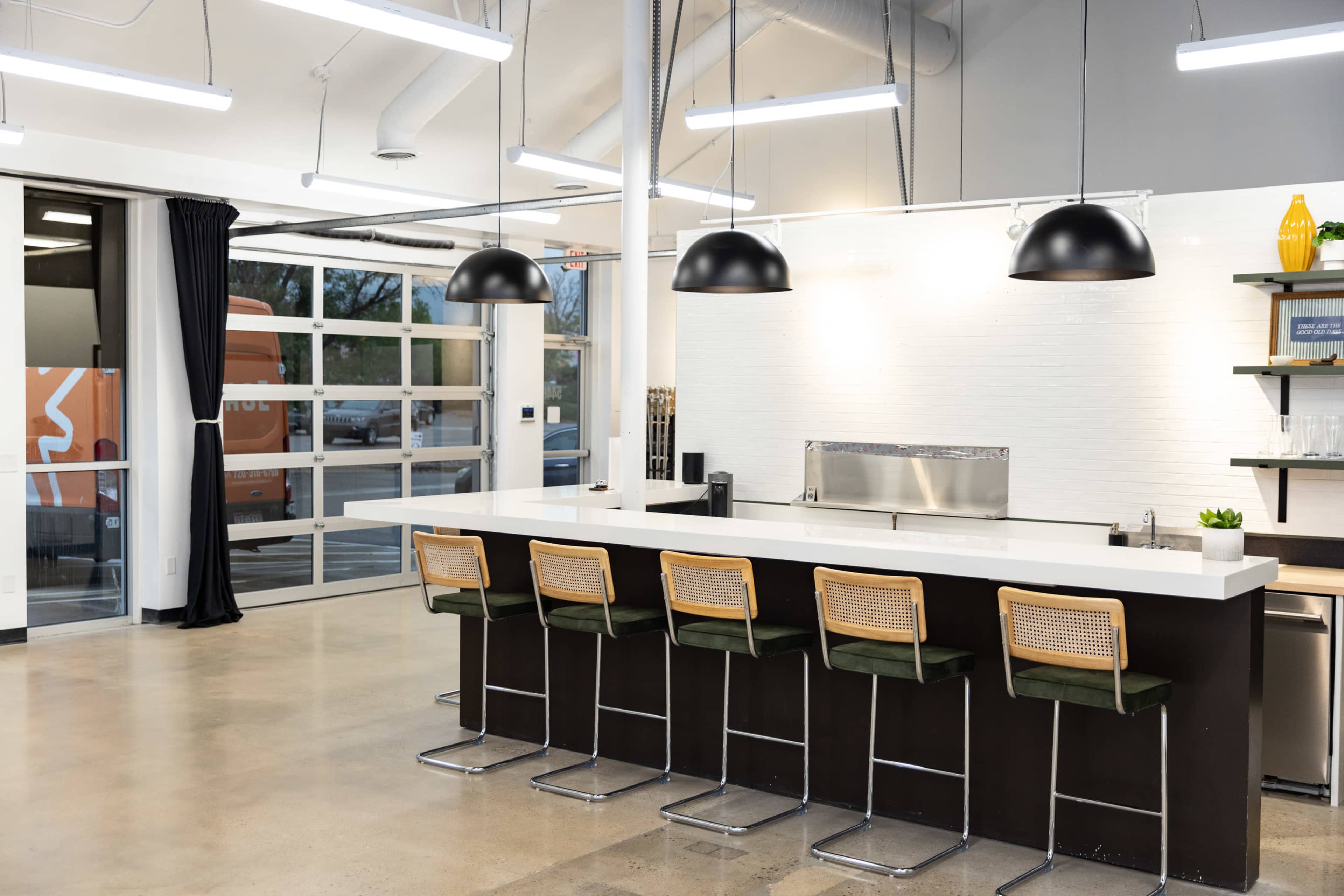 A modern kitchen features a long countertop with six bar stools, pendant lights overhead, and a minimalist design.