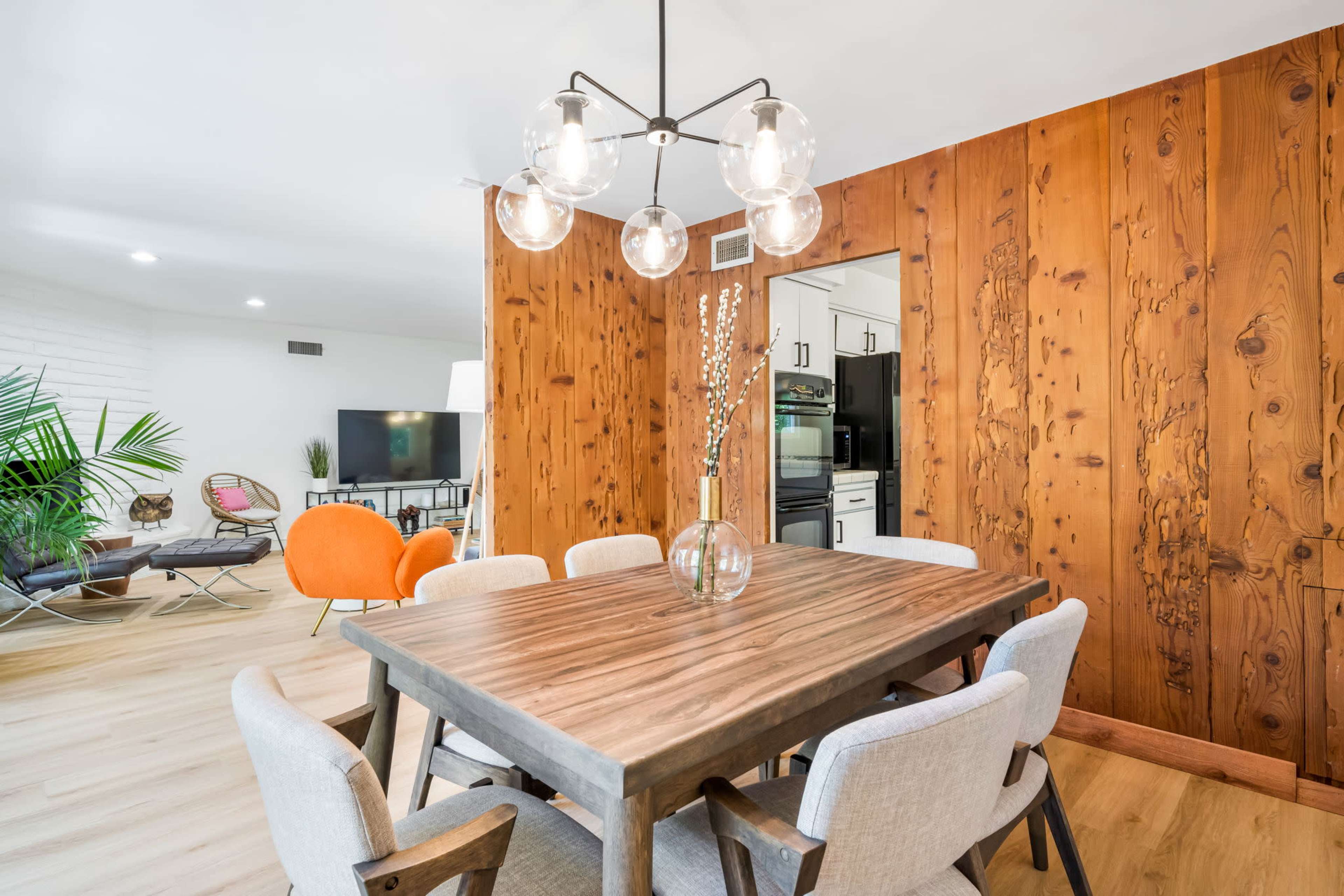 A dining area features a wooden table surrounded by light-colored chairs, with a large chandelier overhead and a wall of wooden paneling.