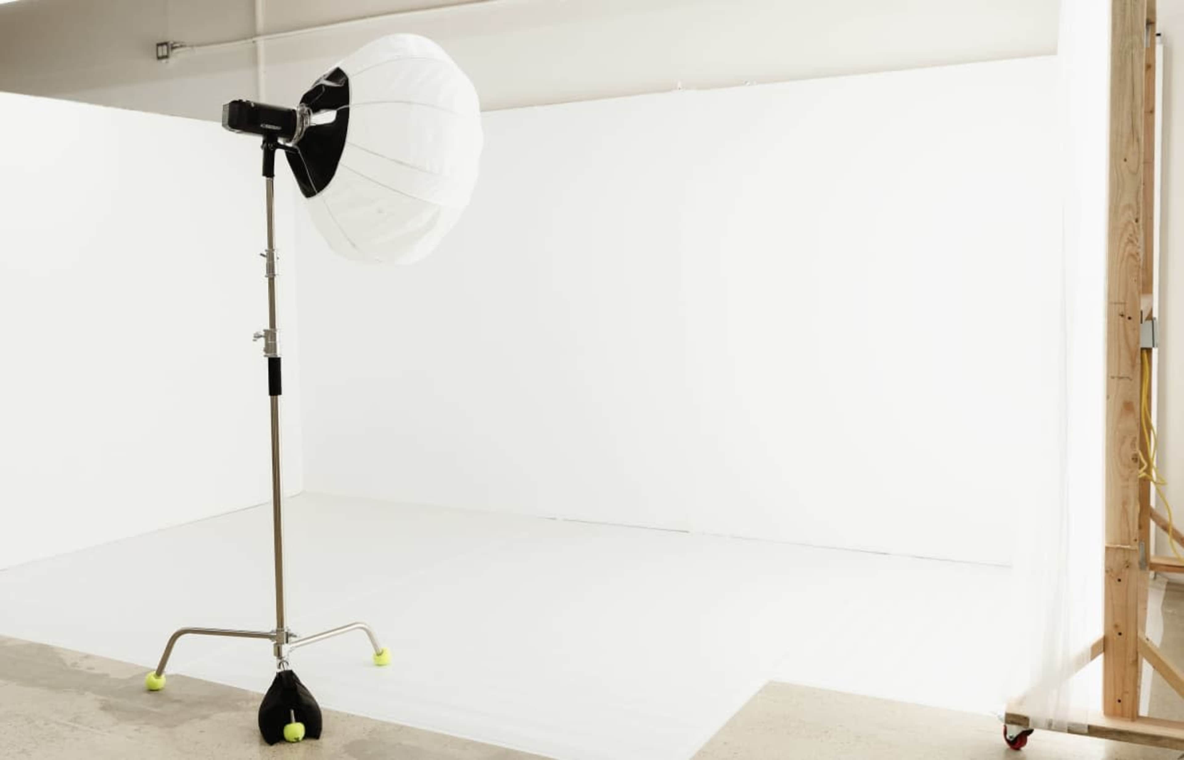 A large lighting setup with a reflective umbrella is positioned in a minimalistic studio space featuring white walls and flooring.