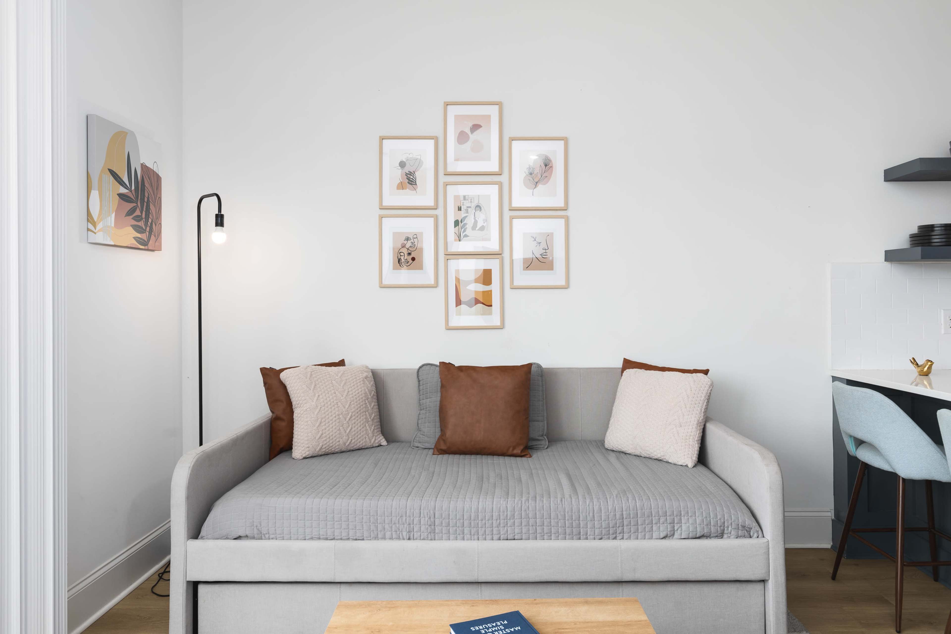 A gray sofa with decorative pillows is positioned in a minimalistic room featuring framed art on the wall and a small dining area with a blue chair.
