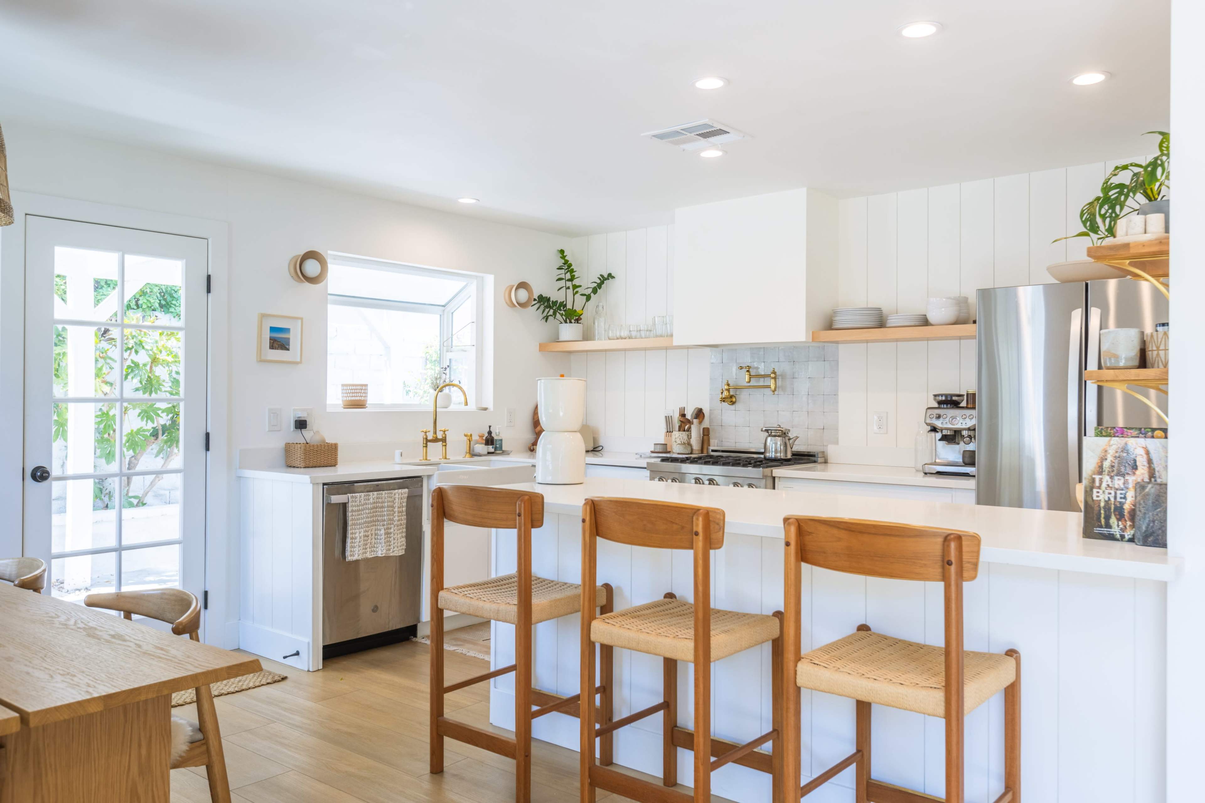 A bright kitchen features a white color scheme with wooden accents, a central island with three barstools, and a window above the sink that provides natural light.