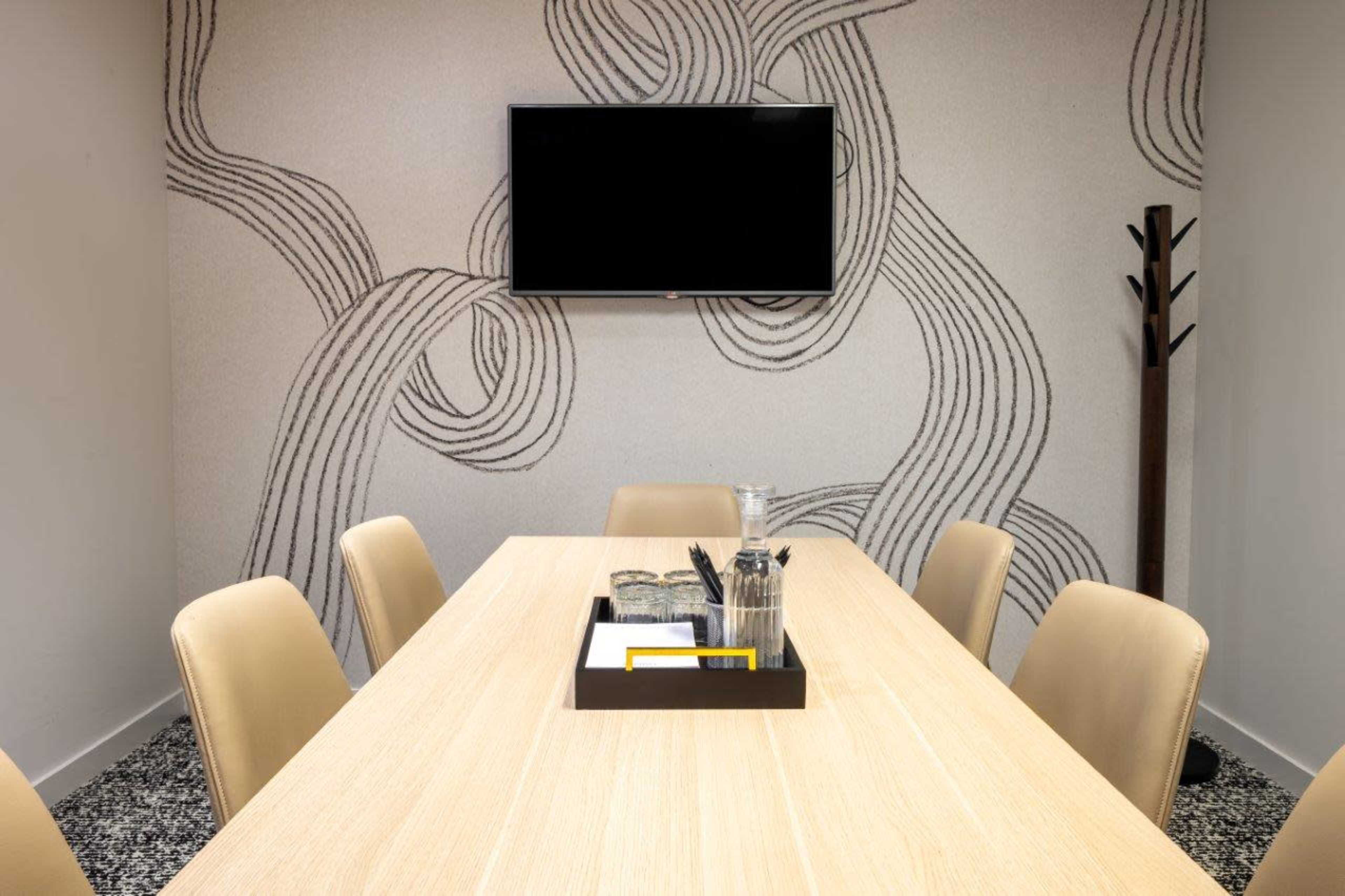 A conference room features a long wooden table surrounded by beige chairs, with a television mounted on a patterned wall and a coat rack in the corner.