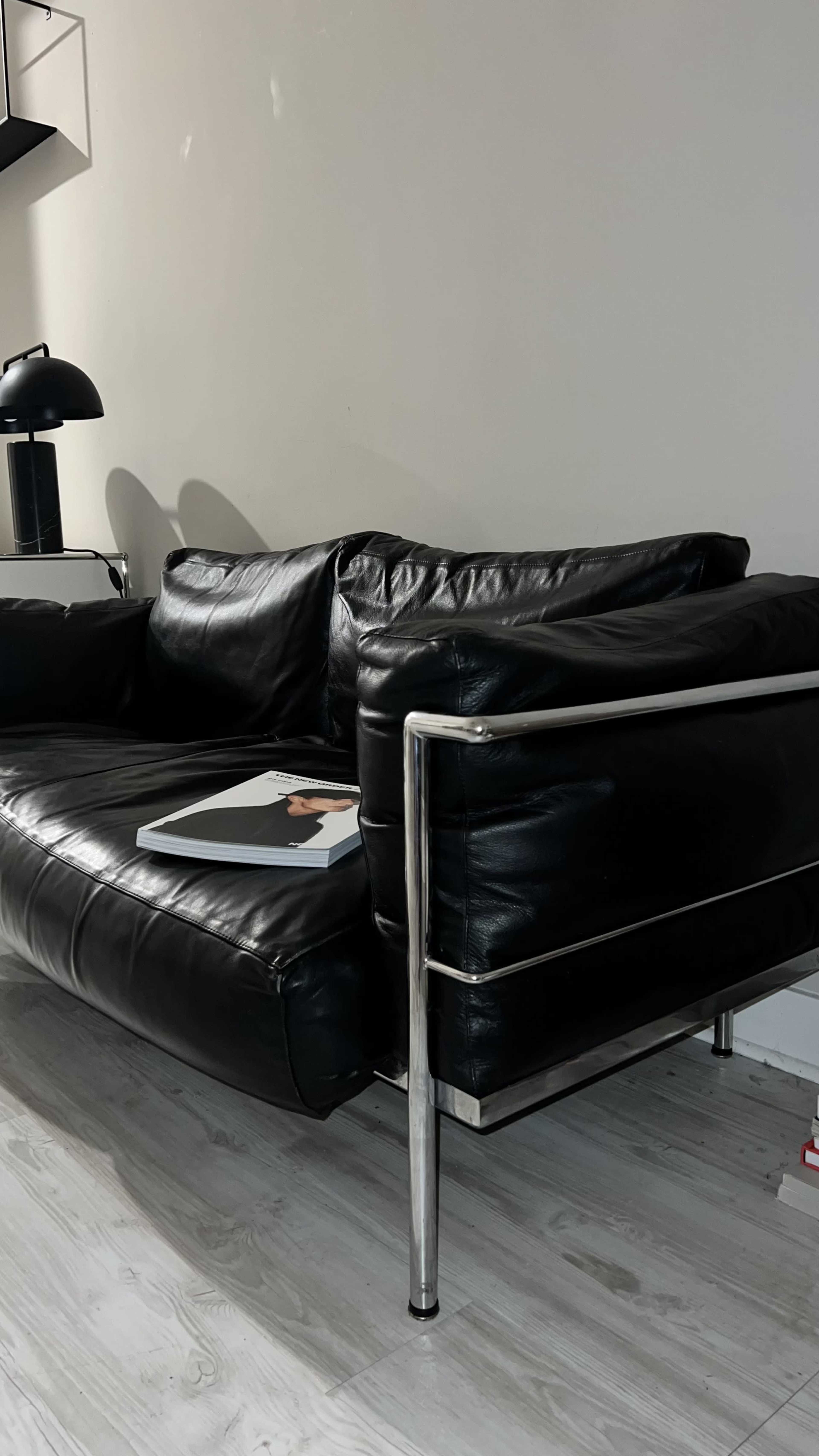 A black leather sofa with a chrome armrest stands next to a modern lamp and a magazine resting on its surface.