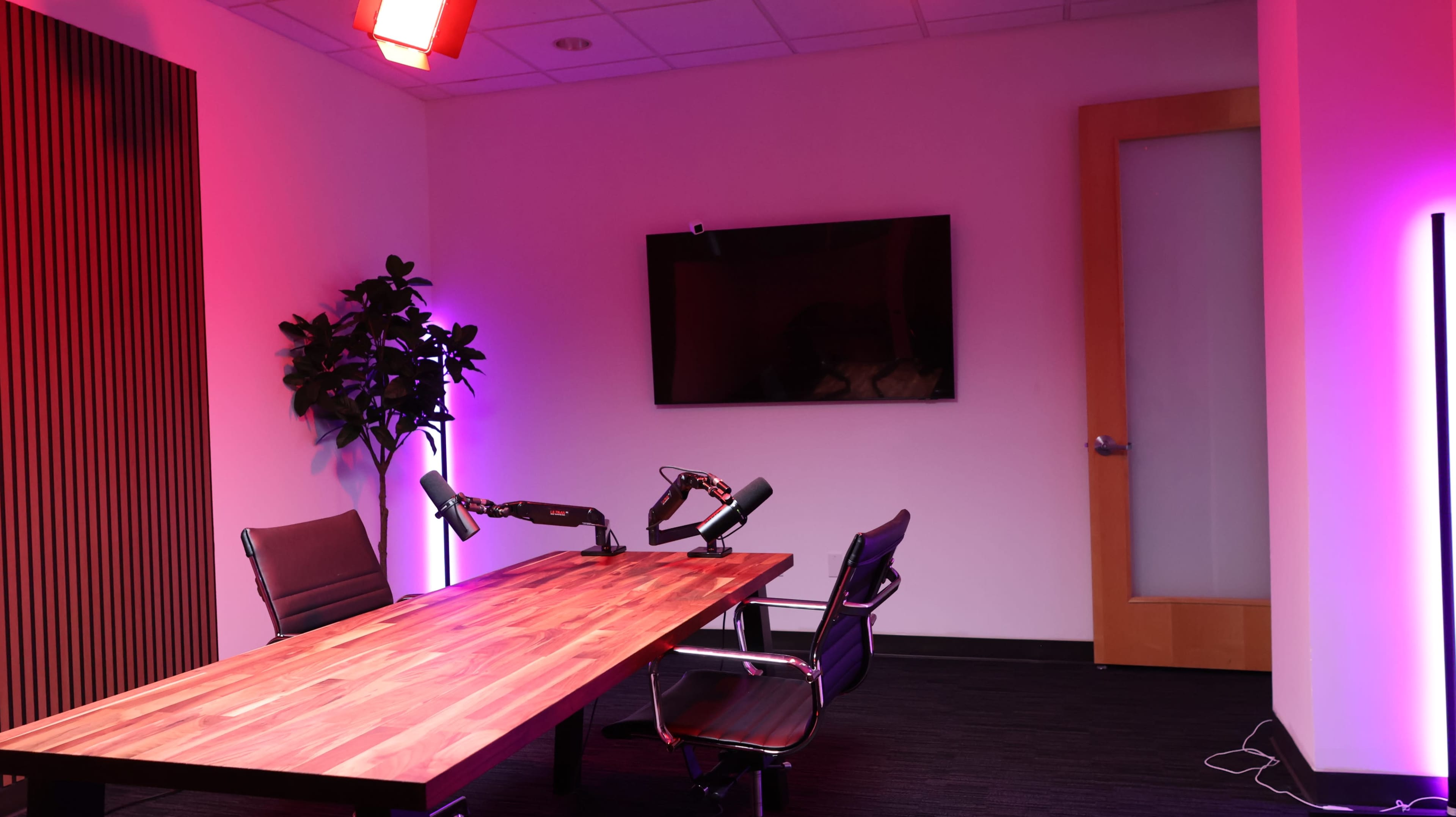 A modern recording studio features a wooden table with two microphones and two chairs, illuminated by colorful lights, and a television mounted on the wall.