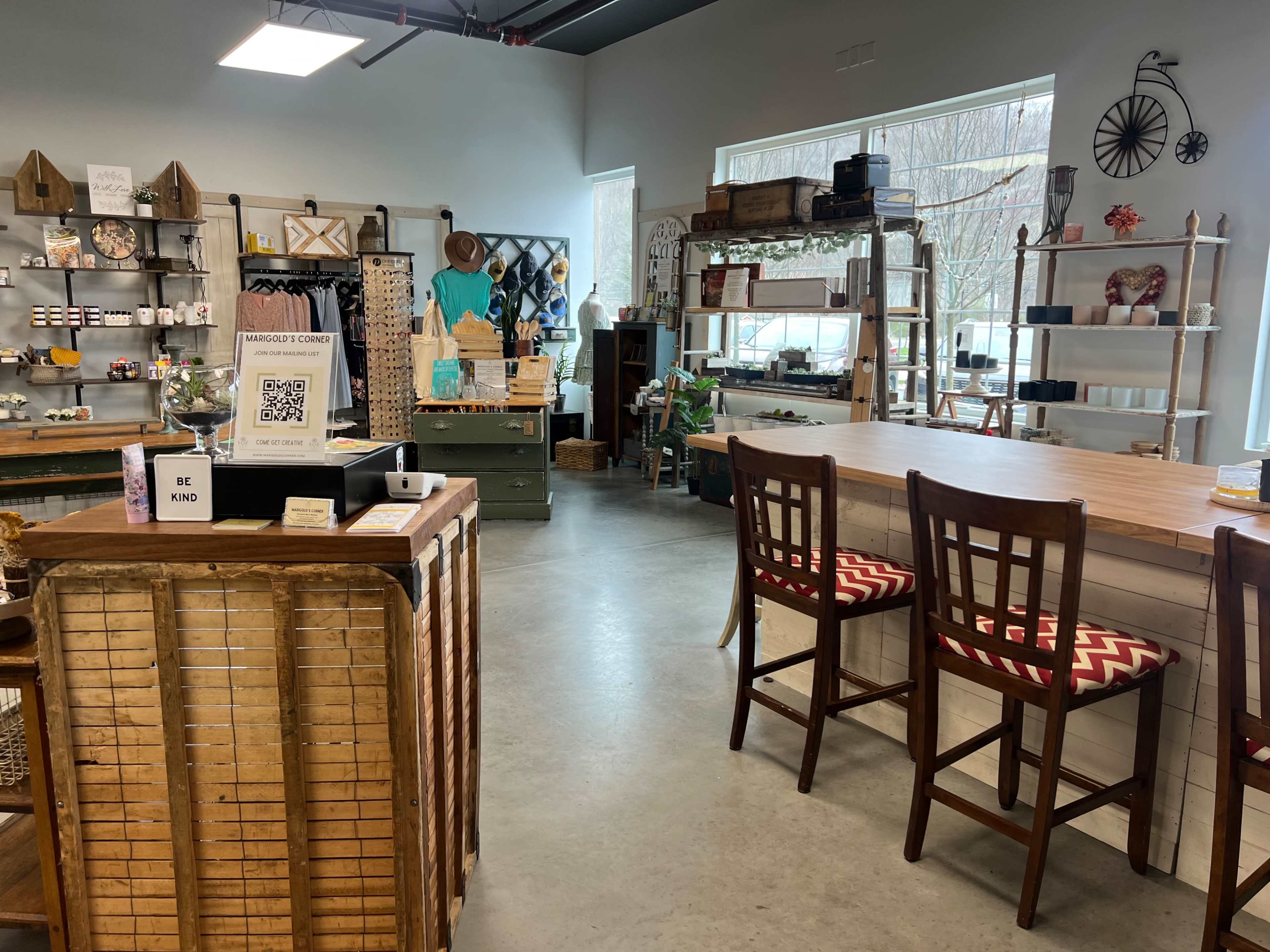 A cozy retail space with wooden furniture, shelves filled with various crafts and home decor items, and a seating area with bar stools.