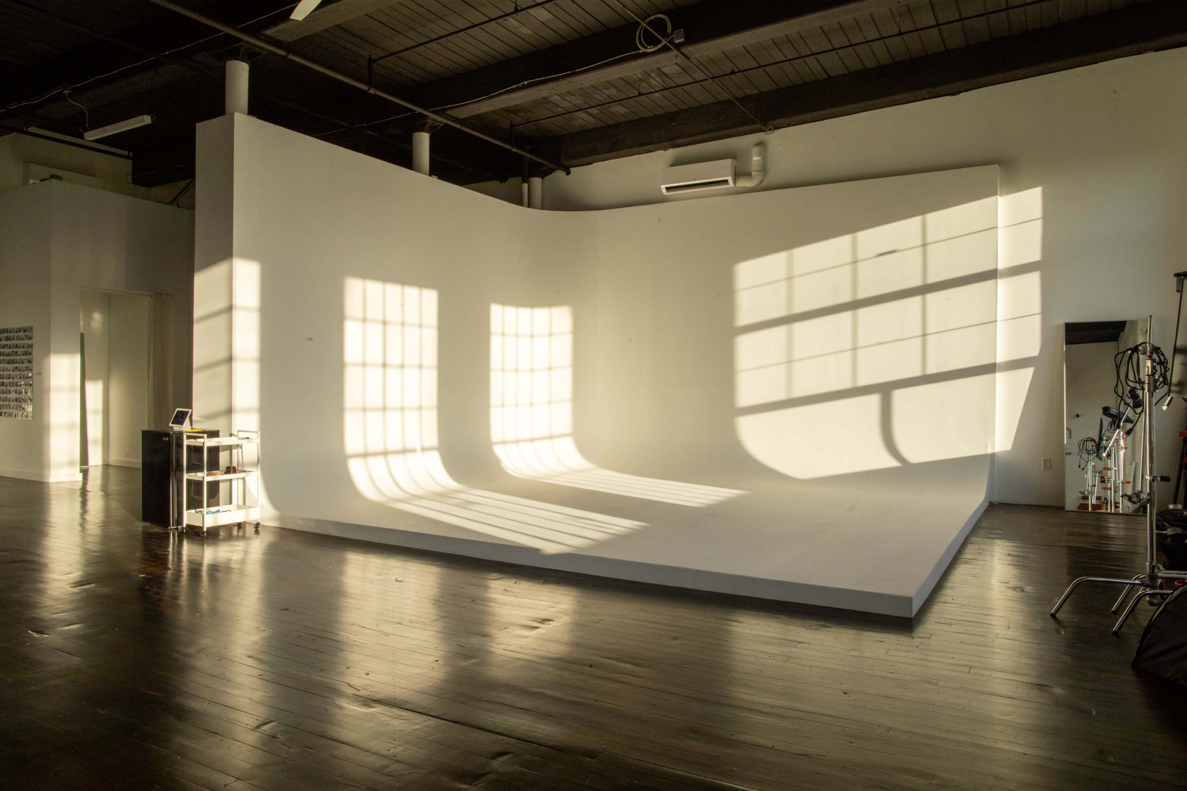 A spacious, empty studio with a curved white backdrop and elongated shadows cast on the floor from large windows.