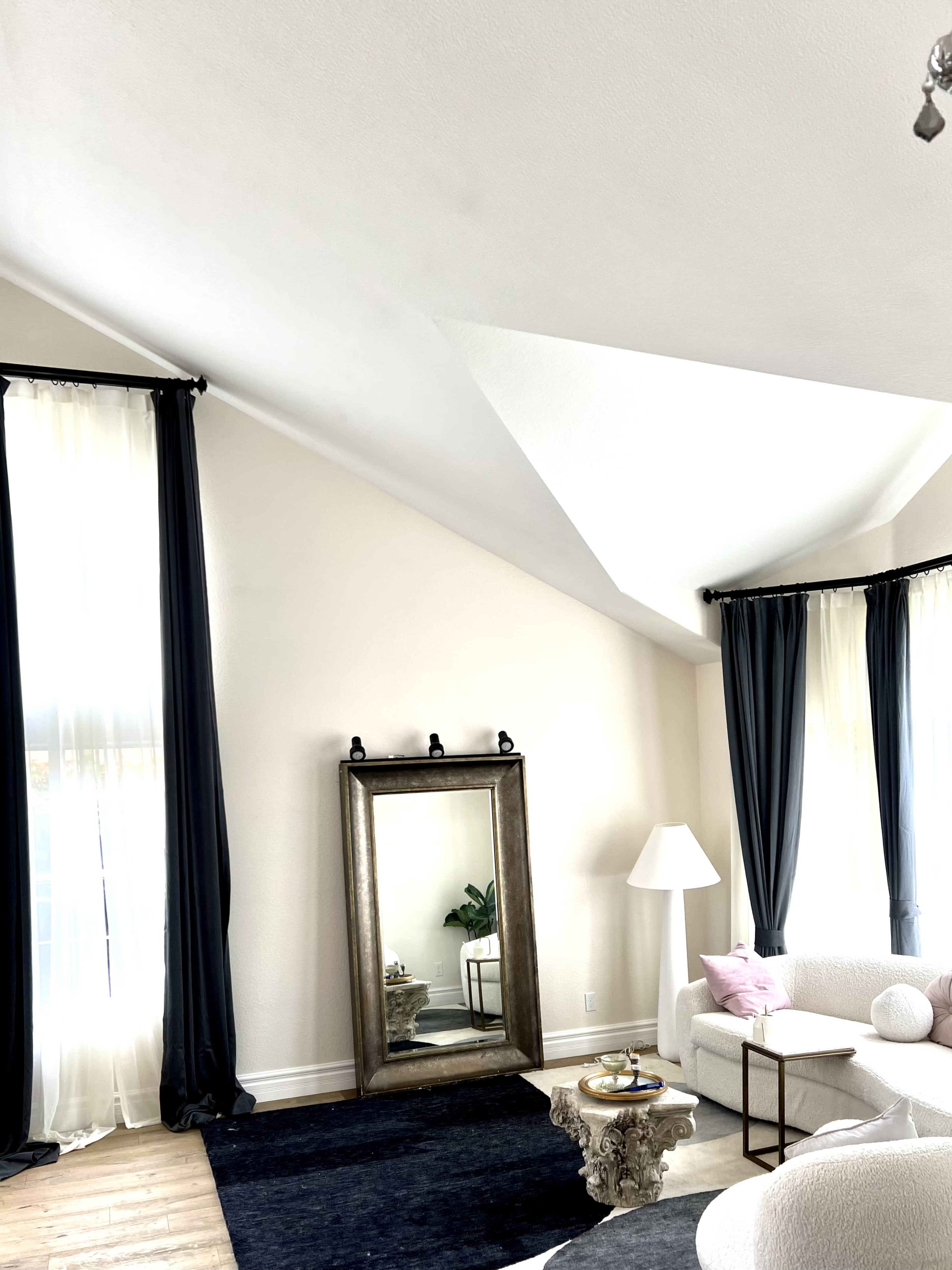 A modern living room features a large mirror, a lamp, and a sofa, with tall windows draped in dark and light curtains against angled ceilings.