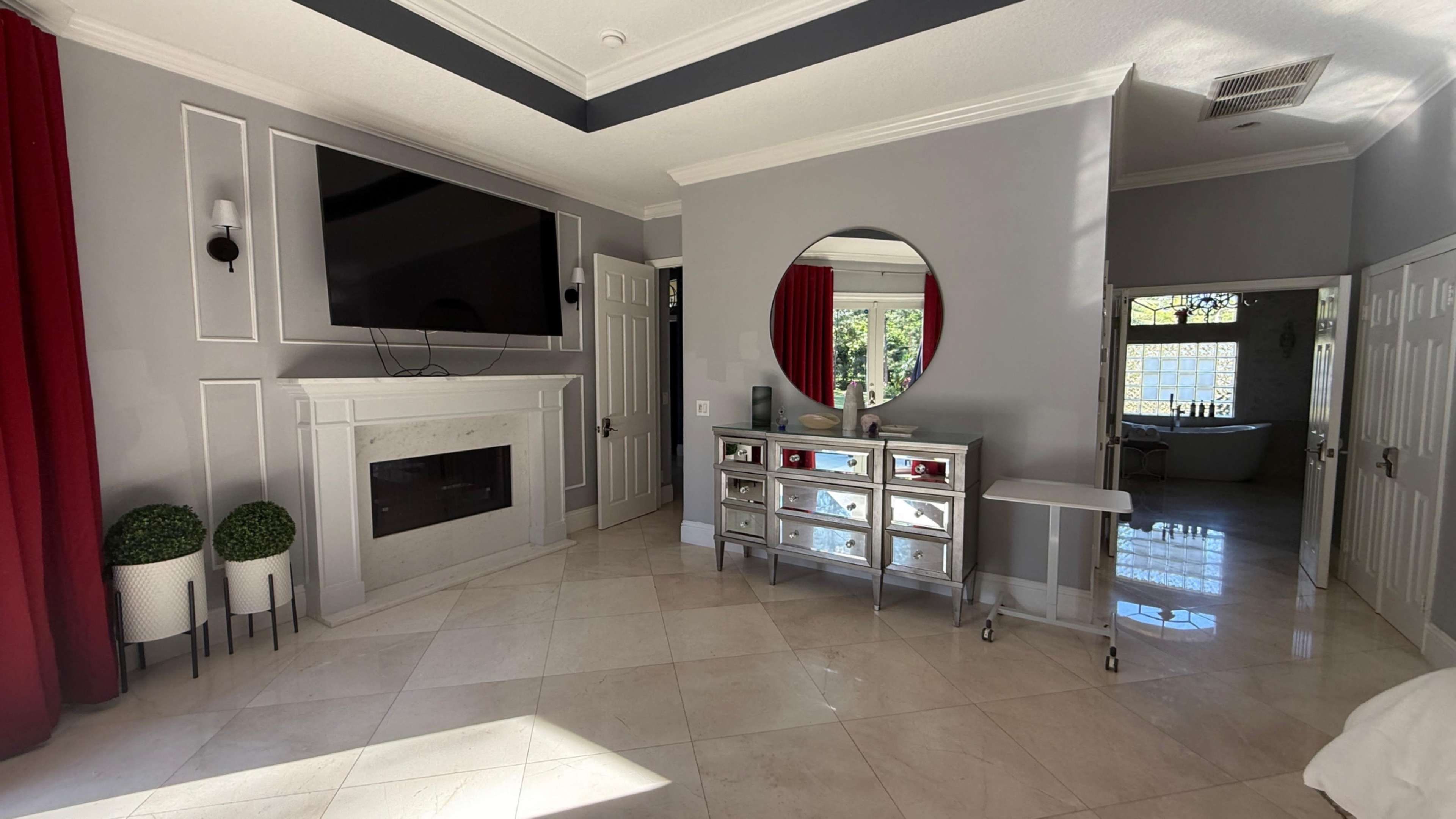 The image features a spacious room with a modern design, showcasing a flat-screen TV mounted above a marble fireplace, a round mirror, a mirrored storage unit, and a doorway leading to another area.