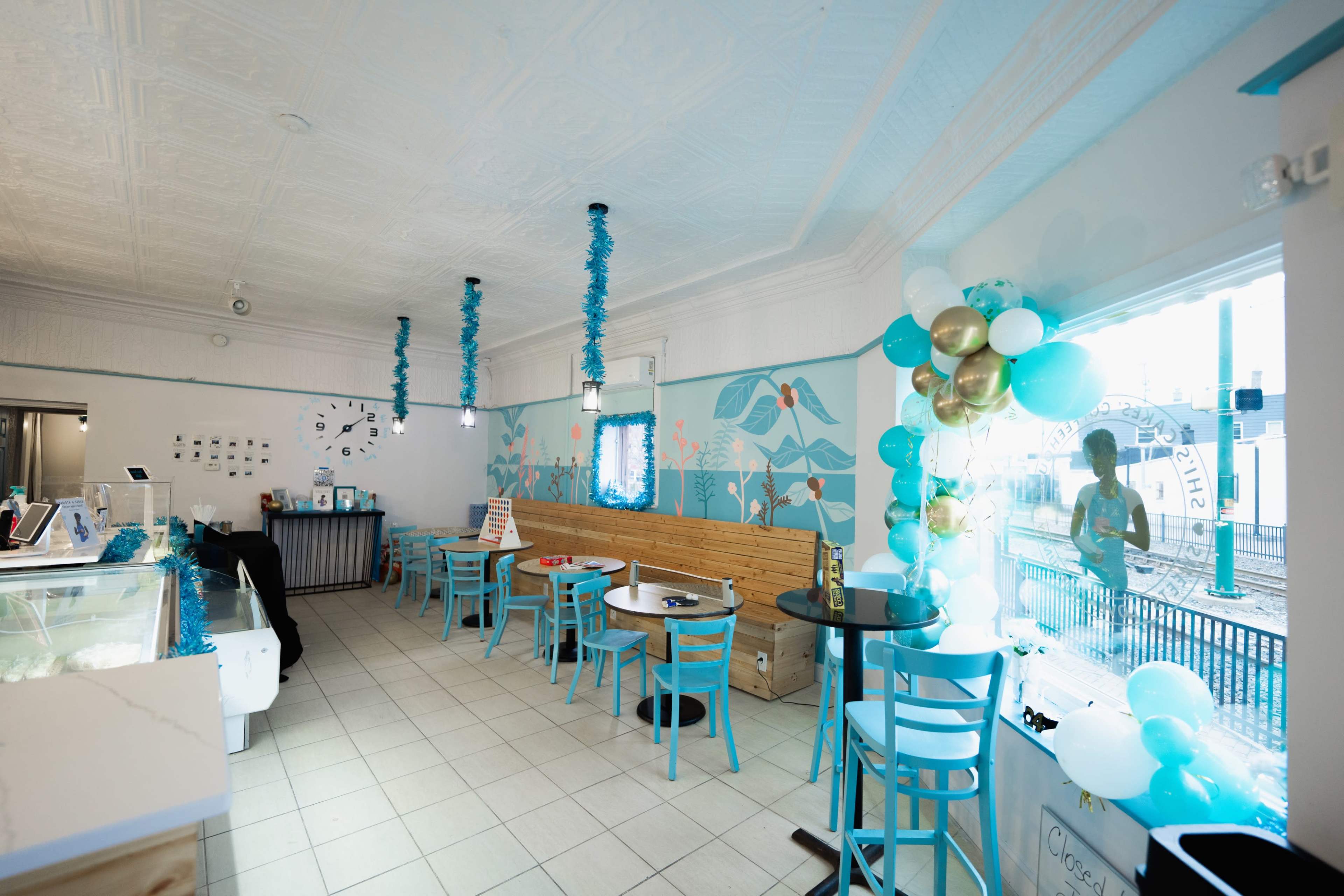 The image depicts a bright, modern ice cream shop with blue and white decor, featuring tables and chairs arranged around the space, and a large window displaying colorful balloons.