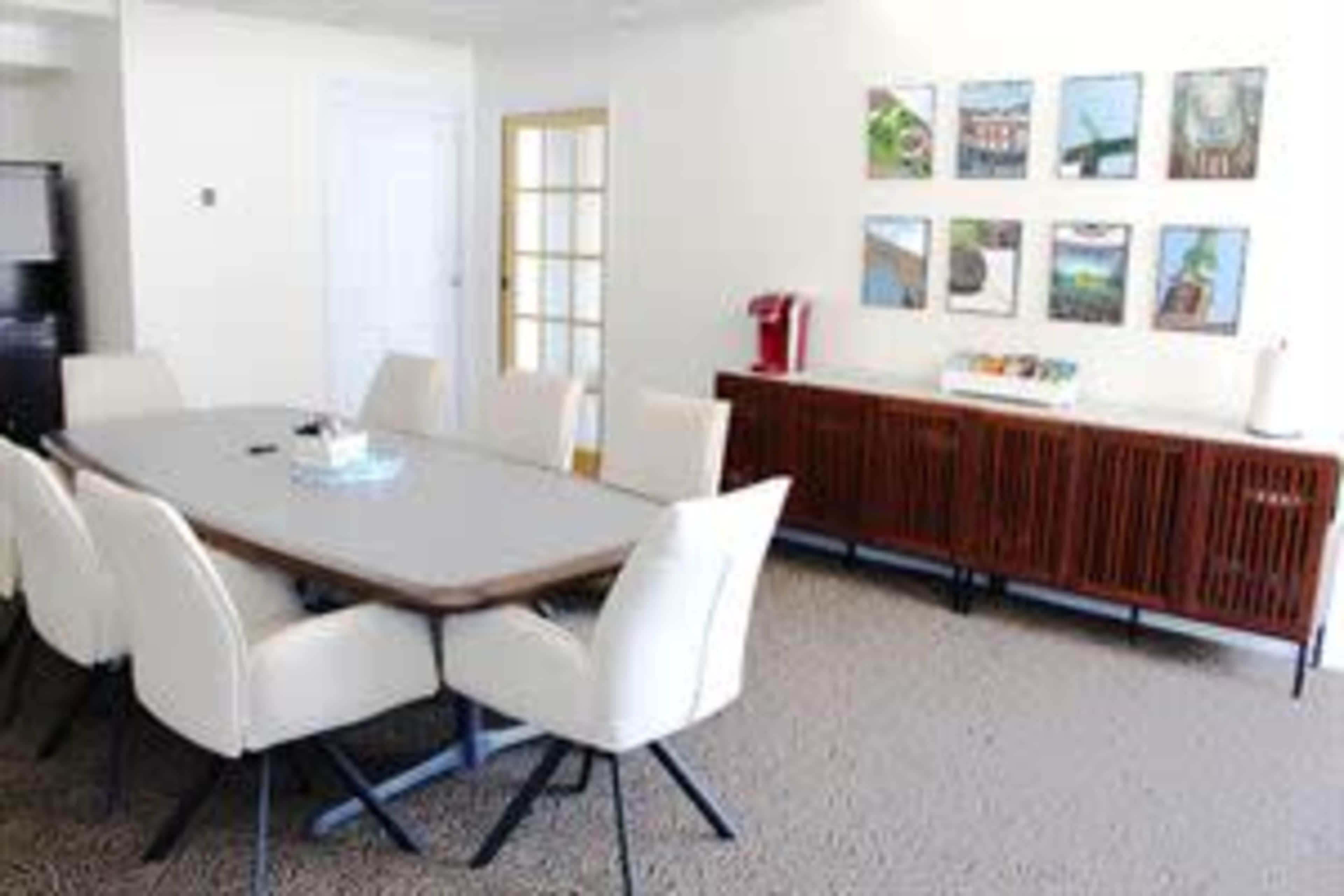 A modern conference room features a long wooden table surrounded by white chairs, with a sideboard and artwork on the wall.