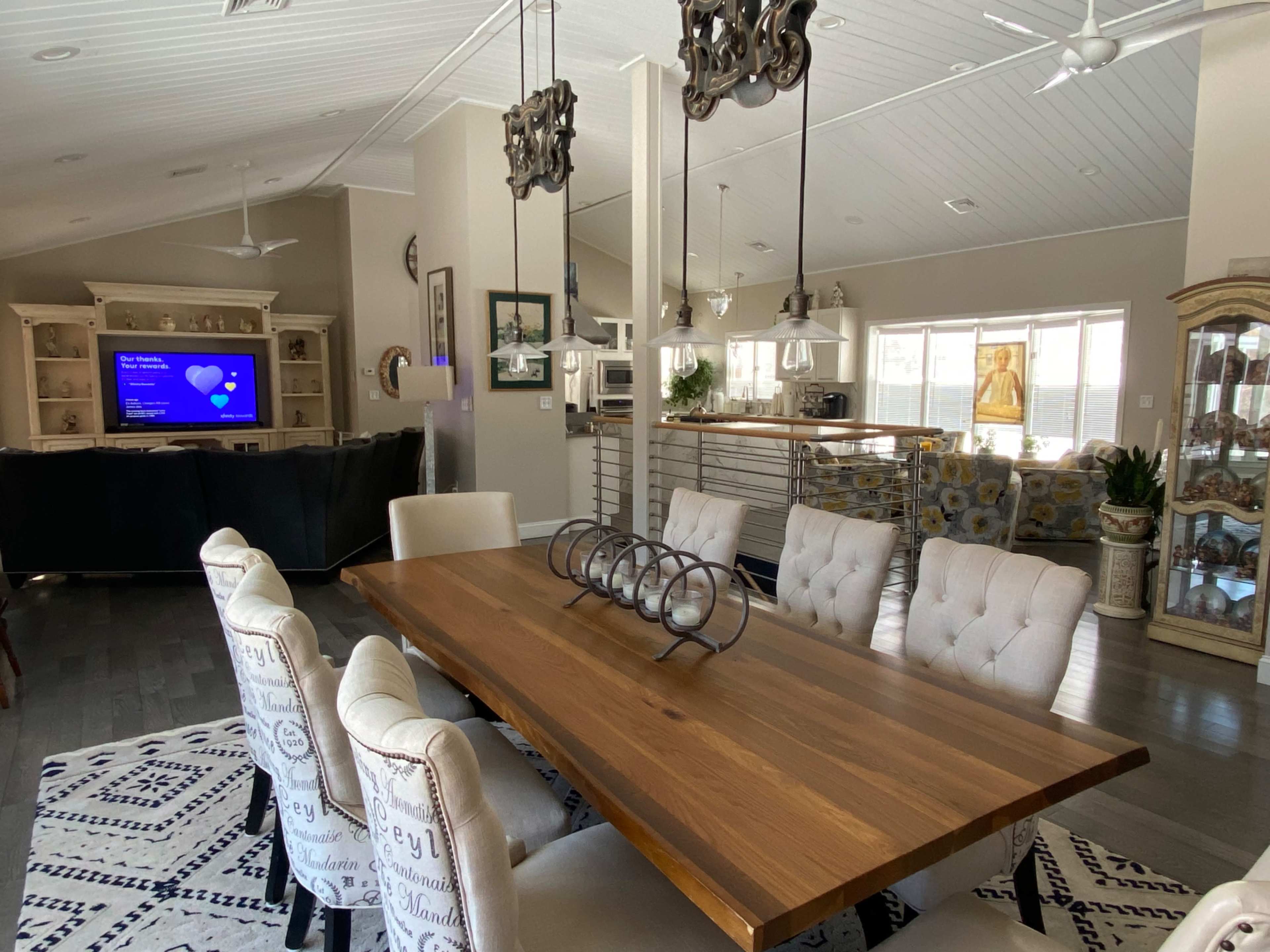 A wooden dining table with upholstered chairs is positioned in a bright, open-concept space featuring a living area and kitchen.