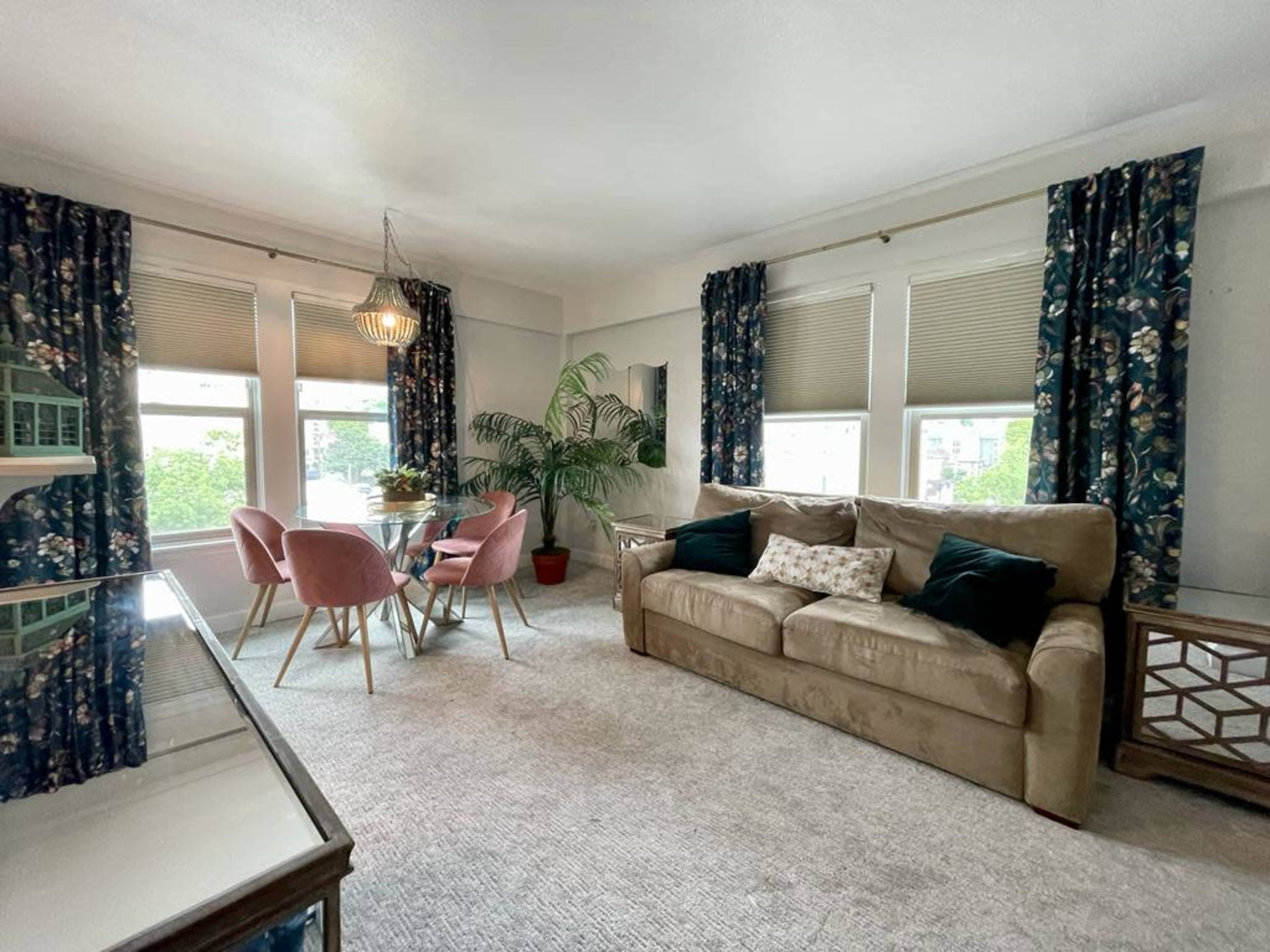 The image shows a living room with a beige couch, a round dining table with pink chairs, a potted plant, and large windows dressed with floral curtains.