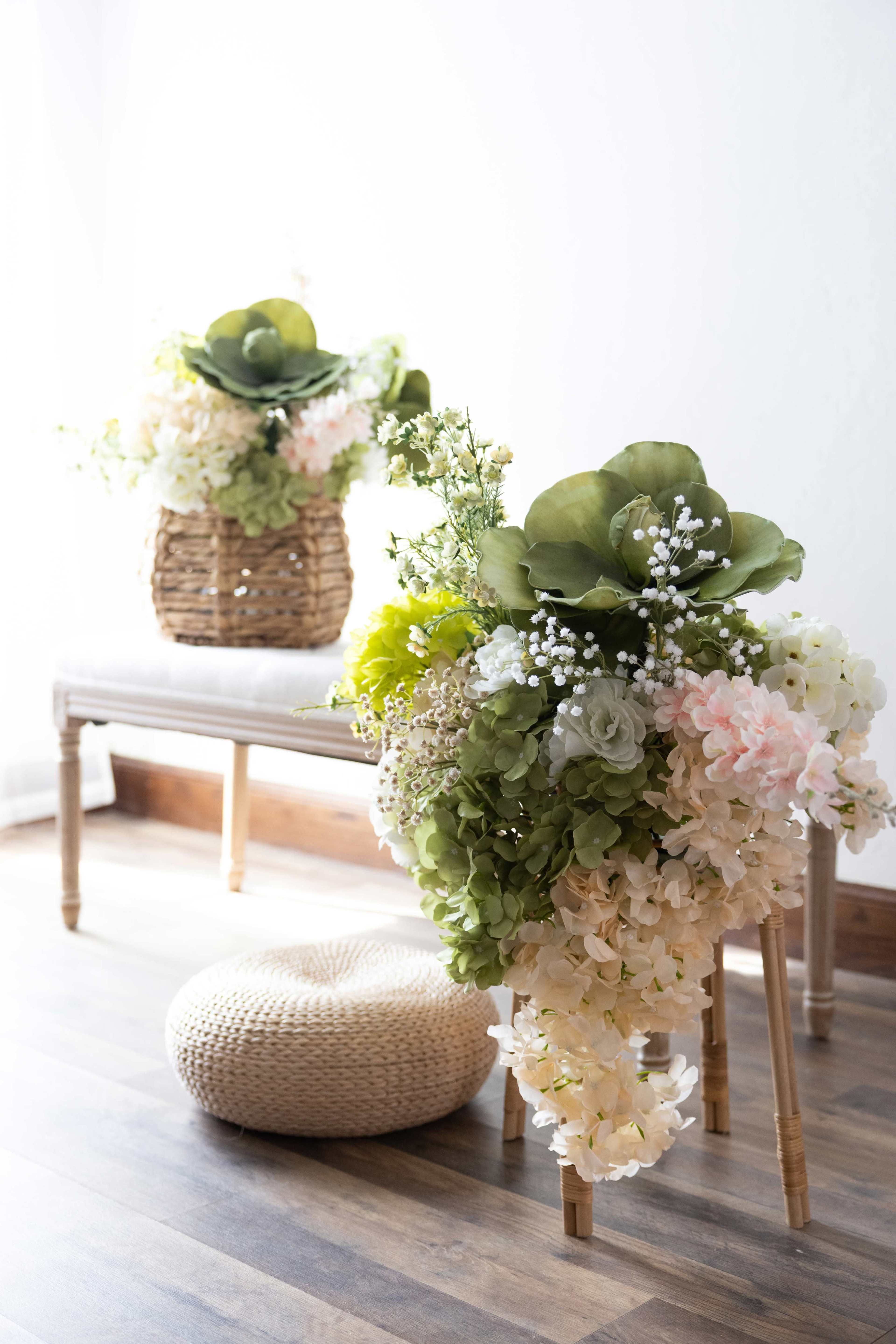 The image features decorative arrangements of flowers in a woven basket and on a small stool, with a round, woven pouf in the foreground.