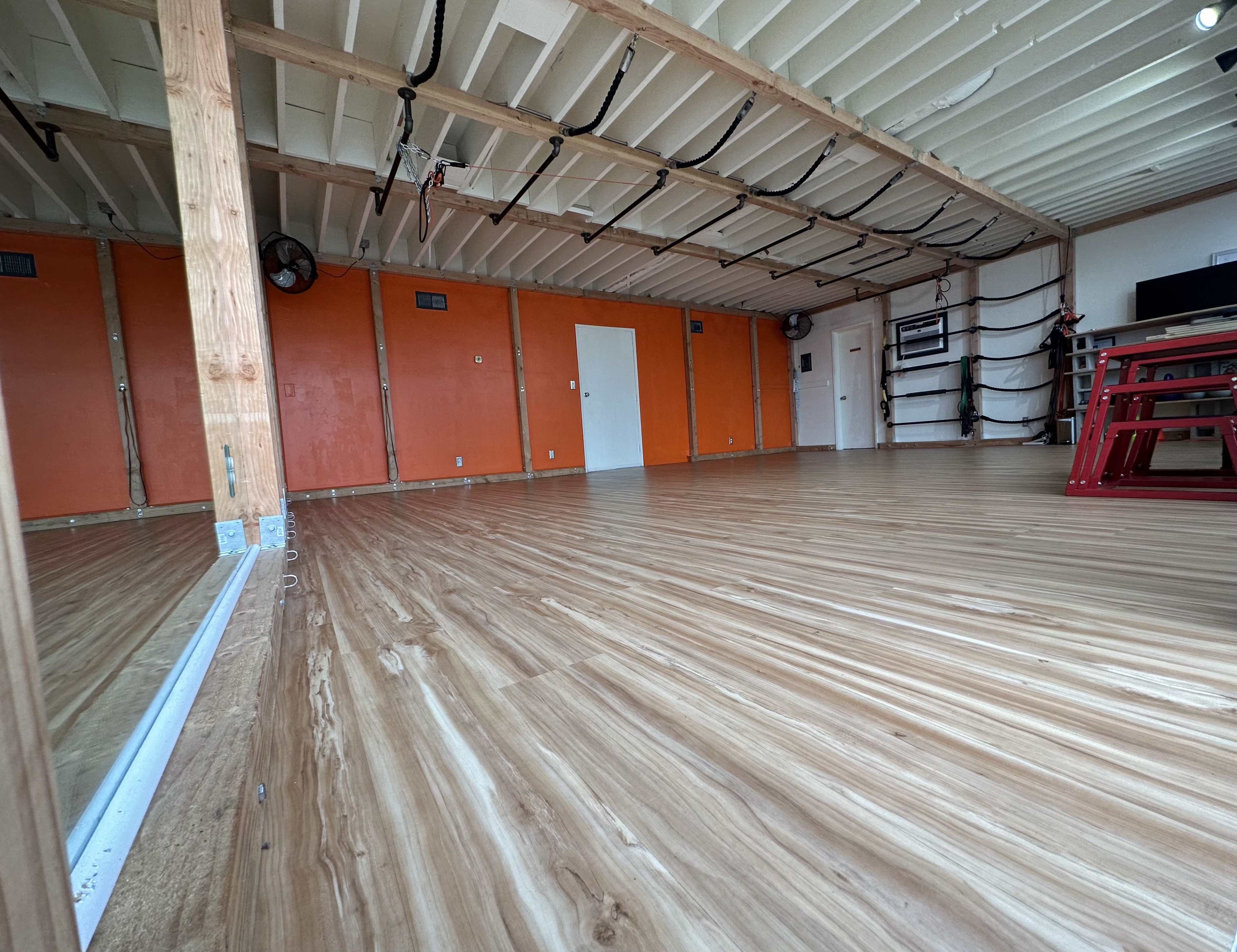 A spacious room with wooden flooring, orange walls, and a ceiling with exposed beams, equipped with sports equipment and storage shelves along one side.
