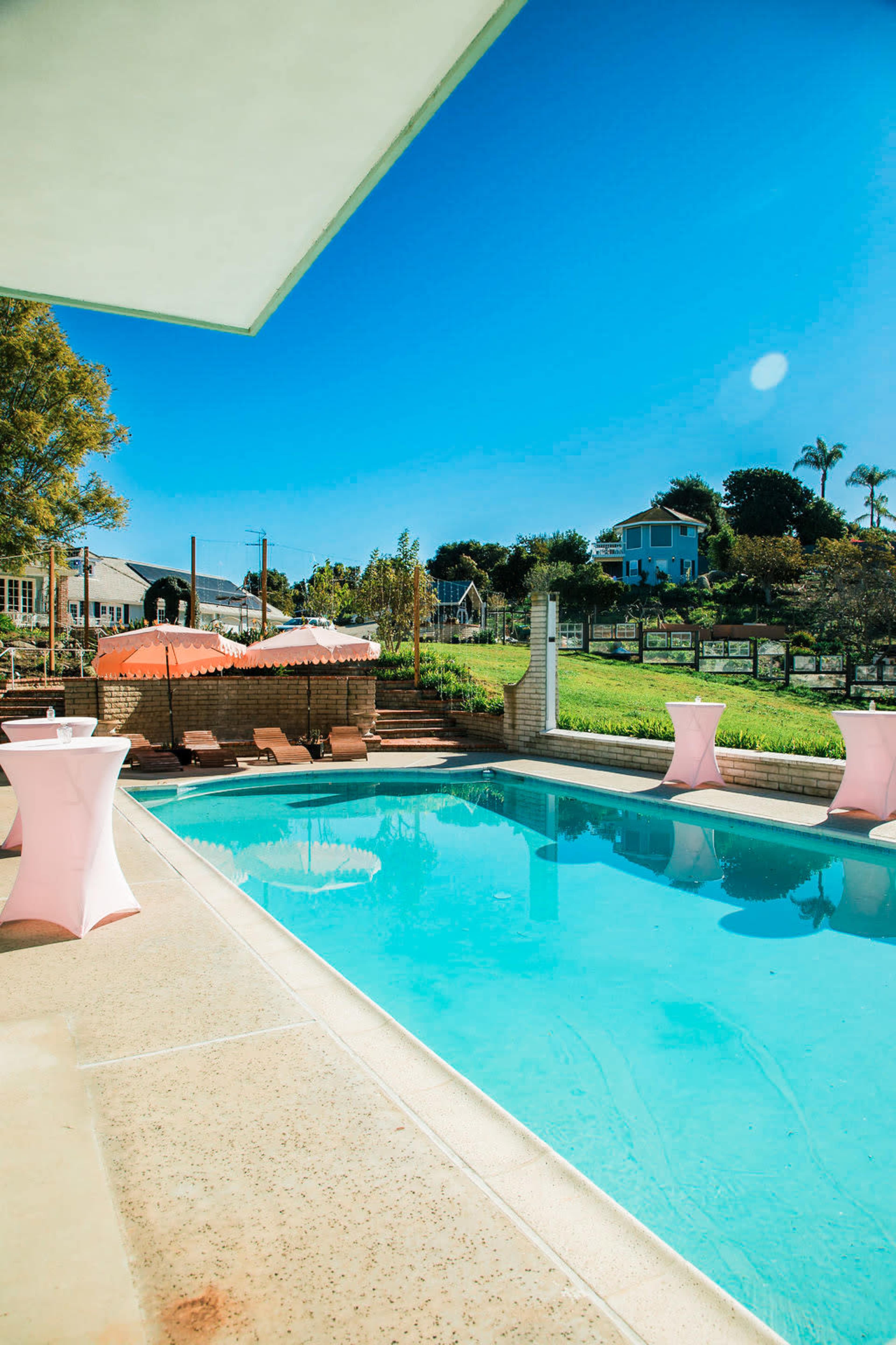 The image shows a clear swimming pool surrounded by pink table settings, with trees and a house in the background under a bright blue sky.