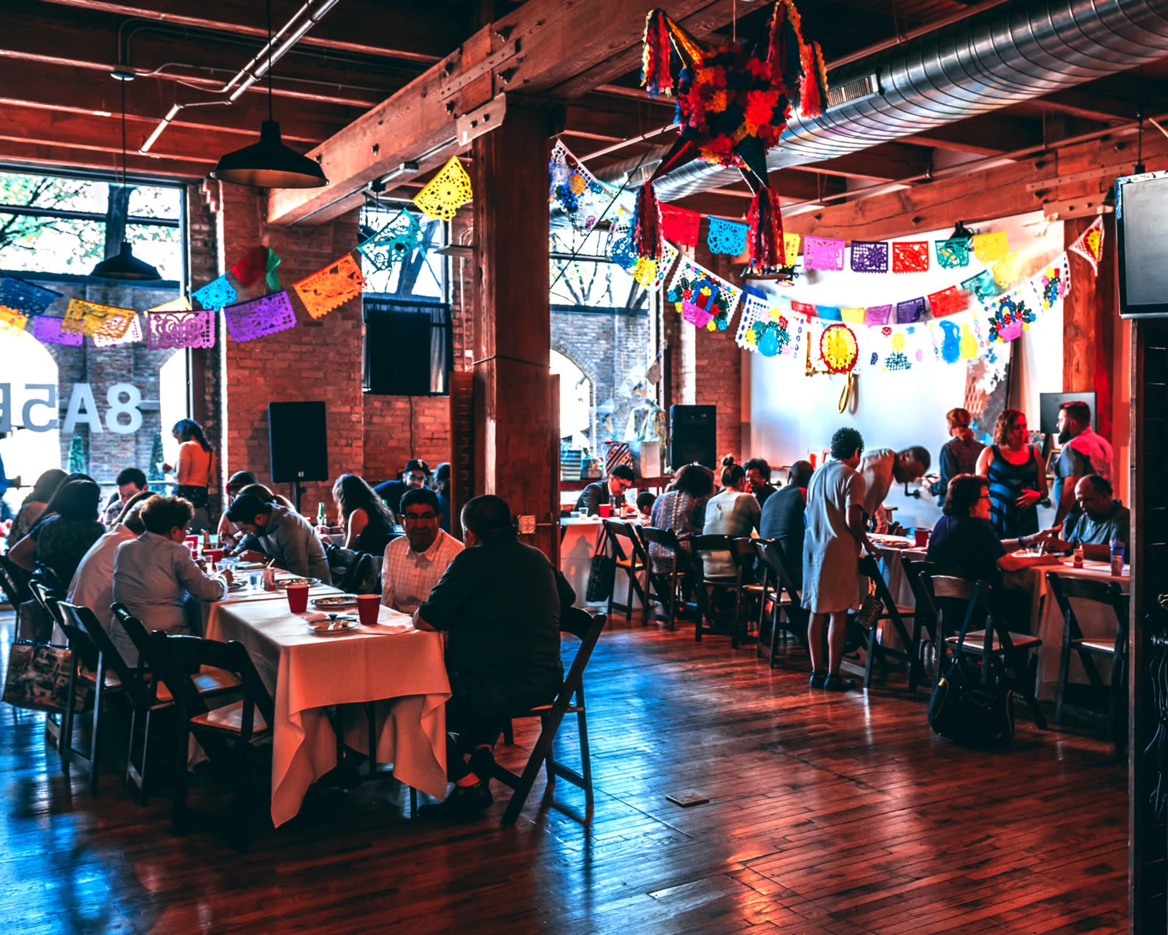 A busy indoor event space features attendees seated at tables, enjoying a meal under colorful decorations and lights.