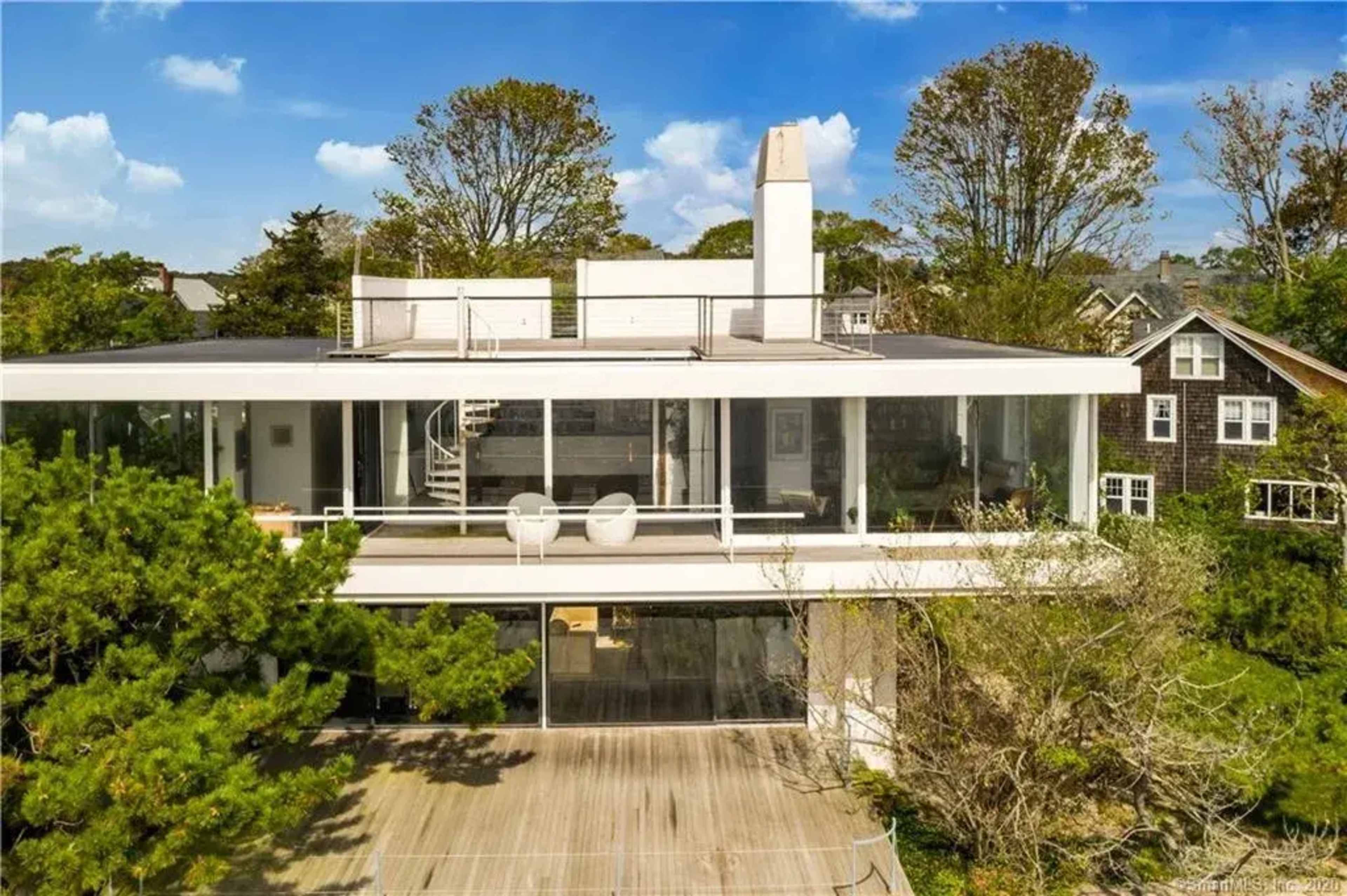 The image shows a modern two-story house with large glass windows surrounded by trees and a wooden deck.