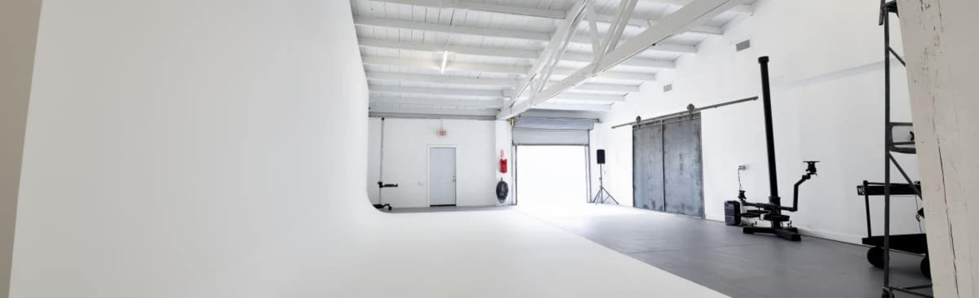 The image shows a spacious, minimalist photography studio with white walls, a smooth floor, and various equipment along one side.