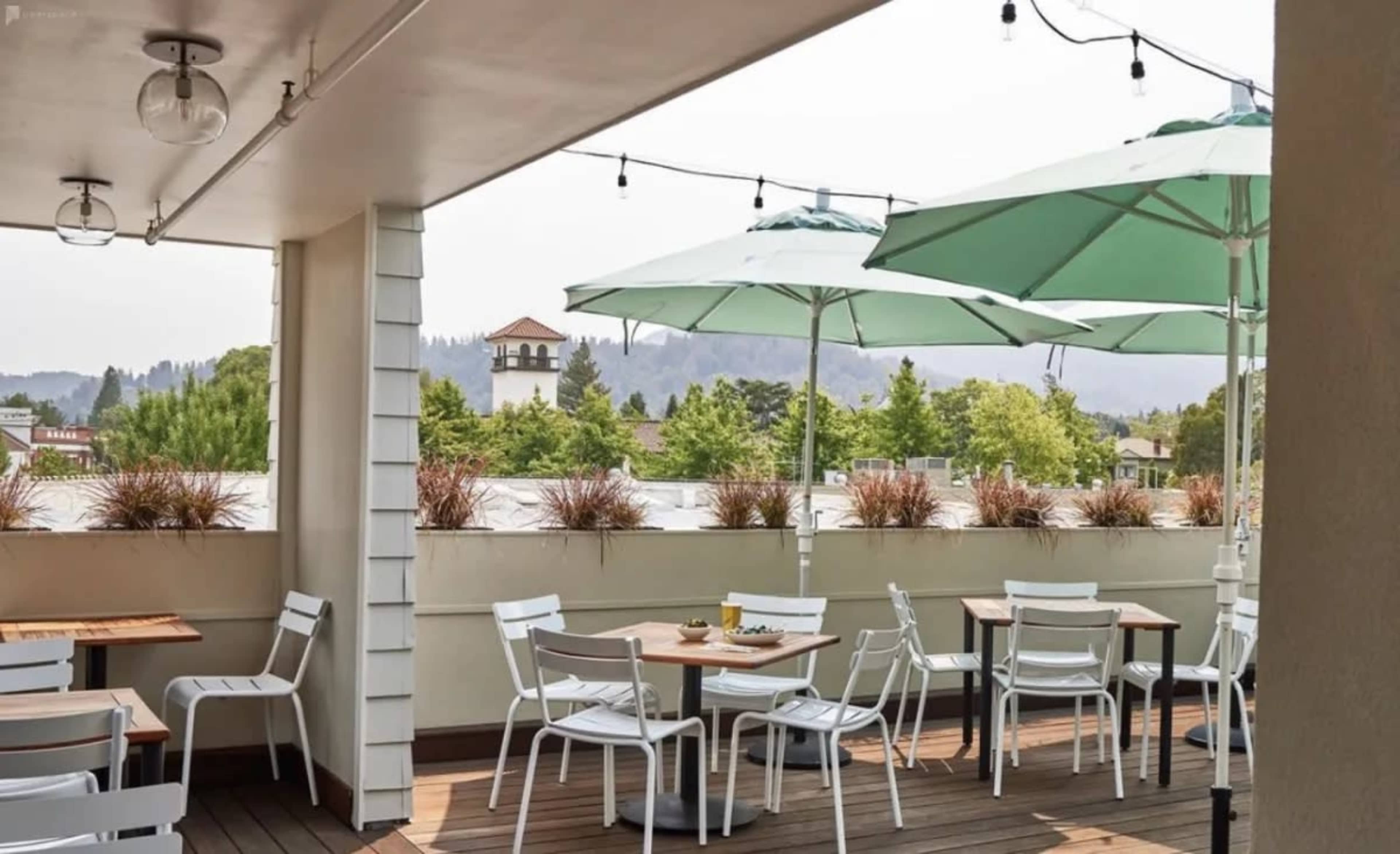 A rooftop patio features white chairs and tables under green umbrellas, surrounded by greenery and distant hills.