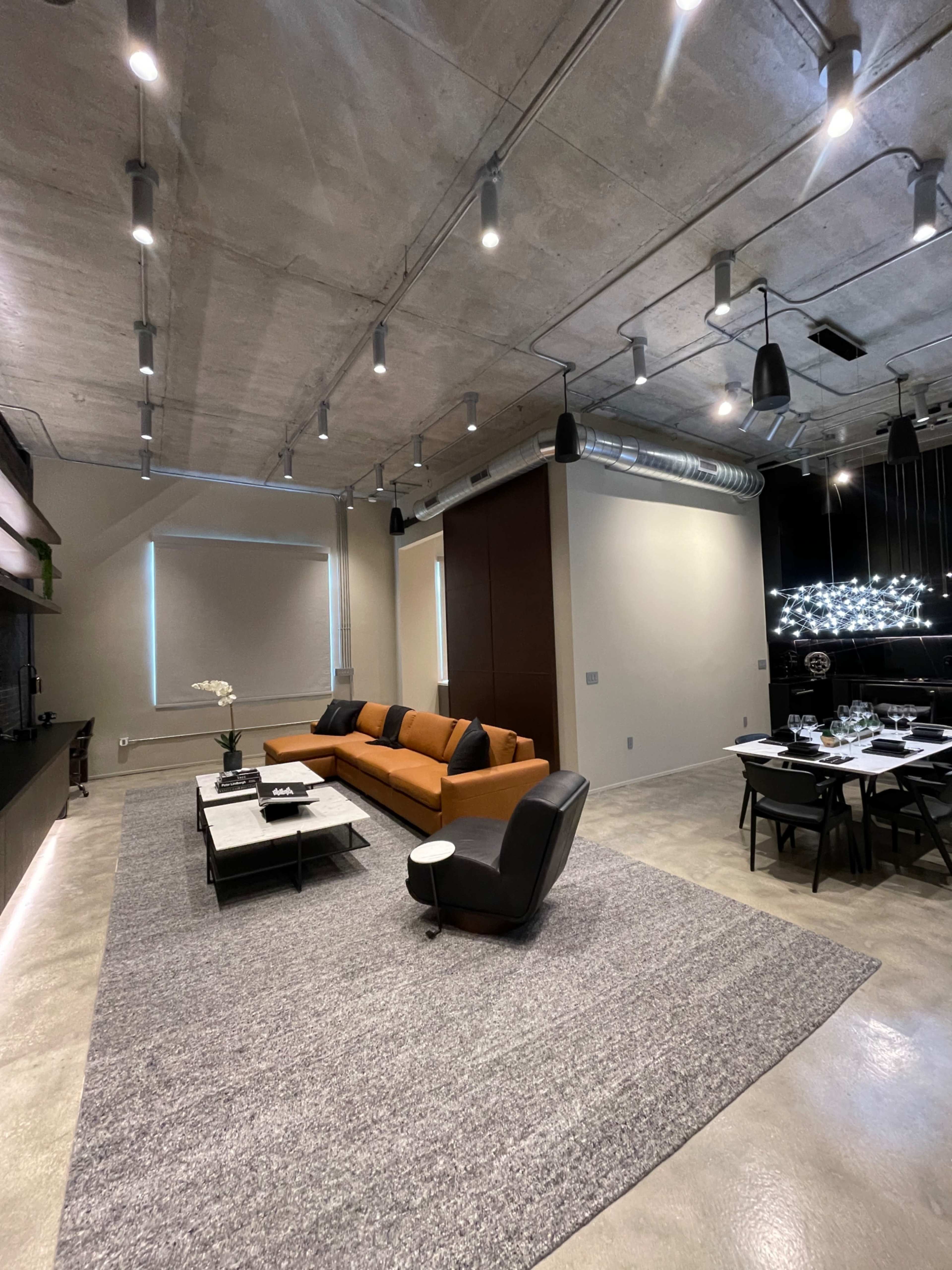 A modern living room with an orange sofa, a black chair, a coffee table, and a dining area illuminated by pendant lights.