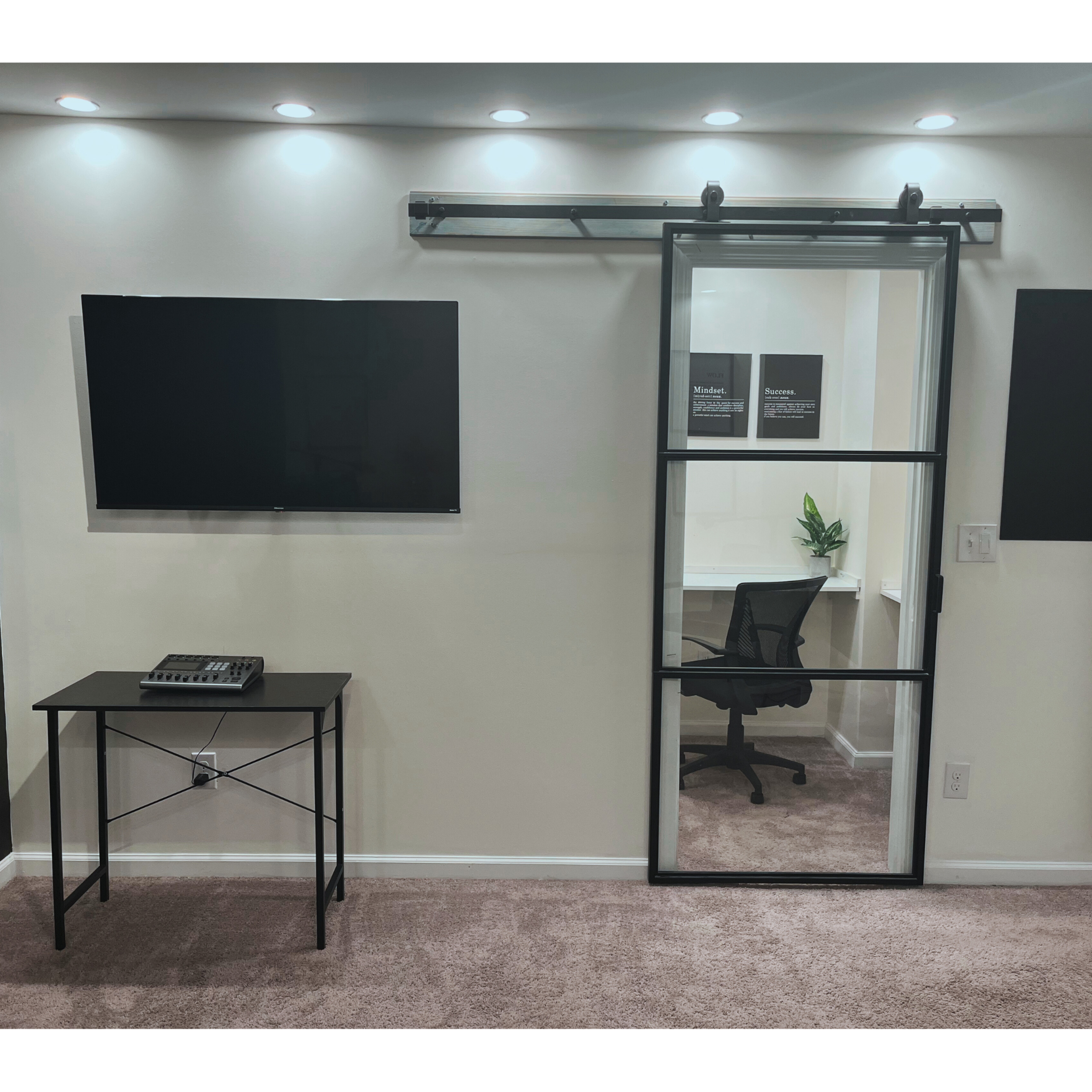 A modern workspace featuring a wall-mounted TV, a black table with equipment, and a sliding glass door leading to an office area with a chair and plant.