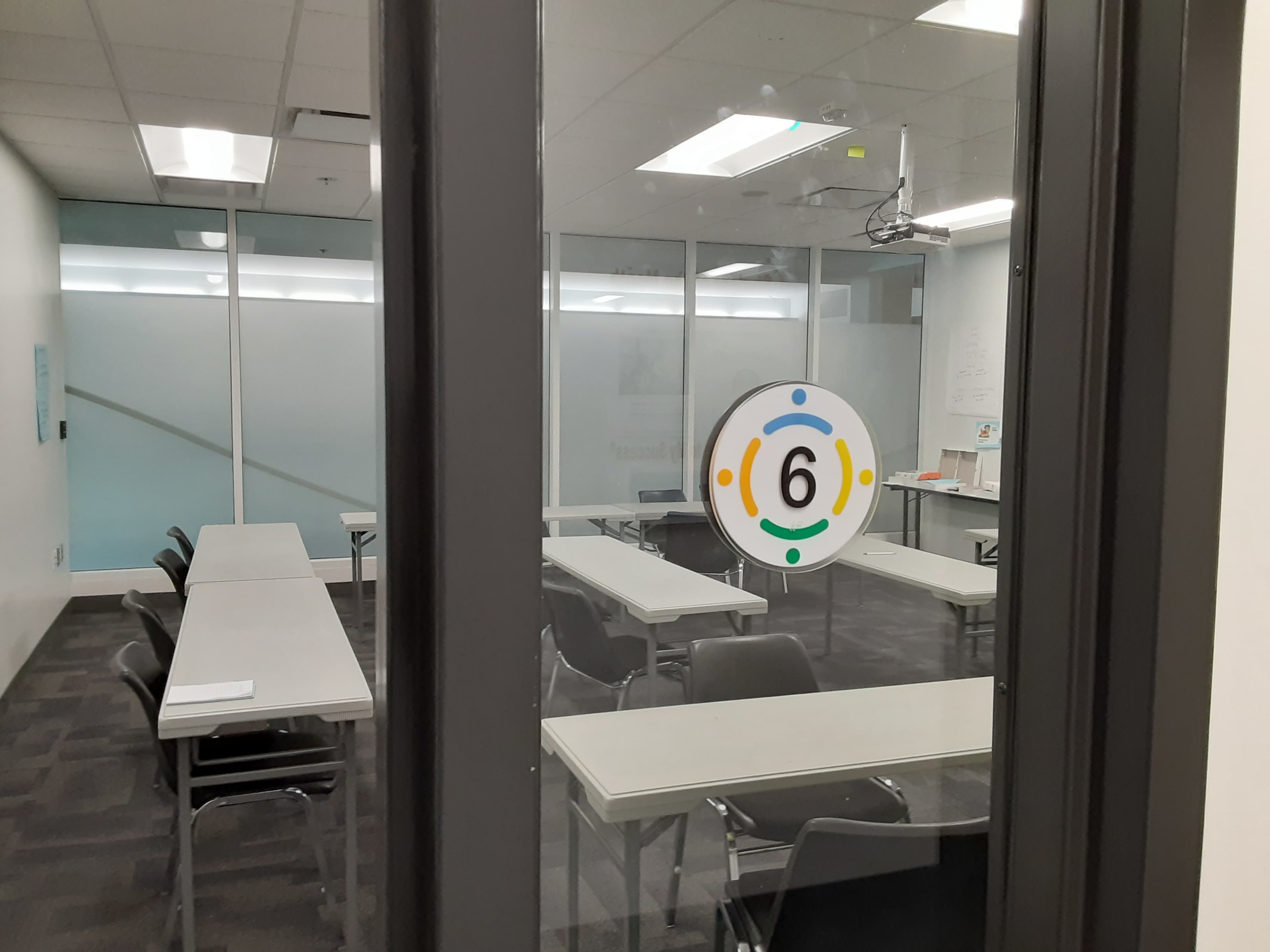 A classroom with several tables and chairs arranged inside, visible through a glass door marked with the number "6."