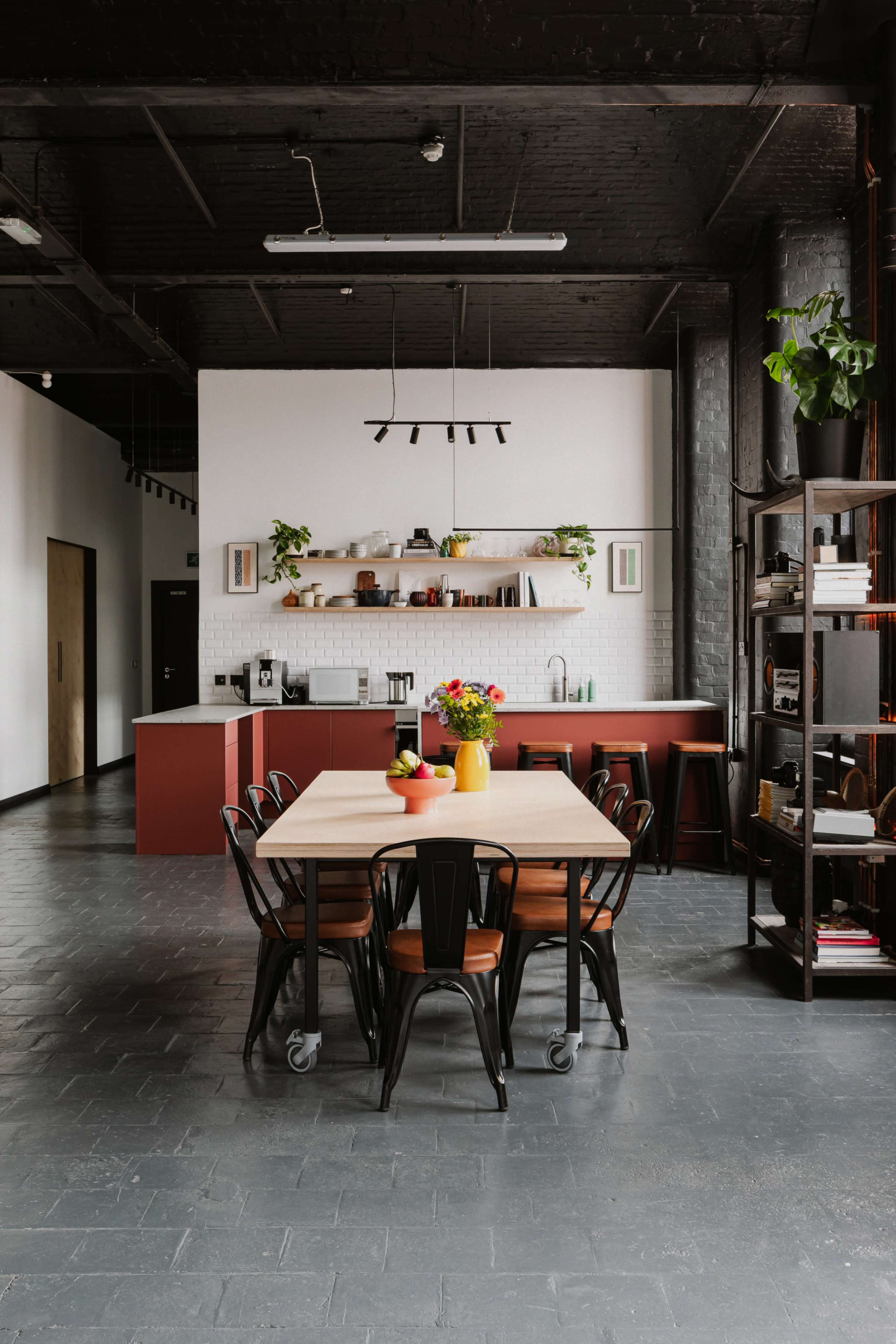 The image shows a modern kitchen and dining area featuring a large wooden table surrounded by black metal chairs, with a shelf displaying kitchen items in the background.