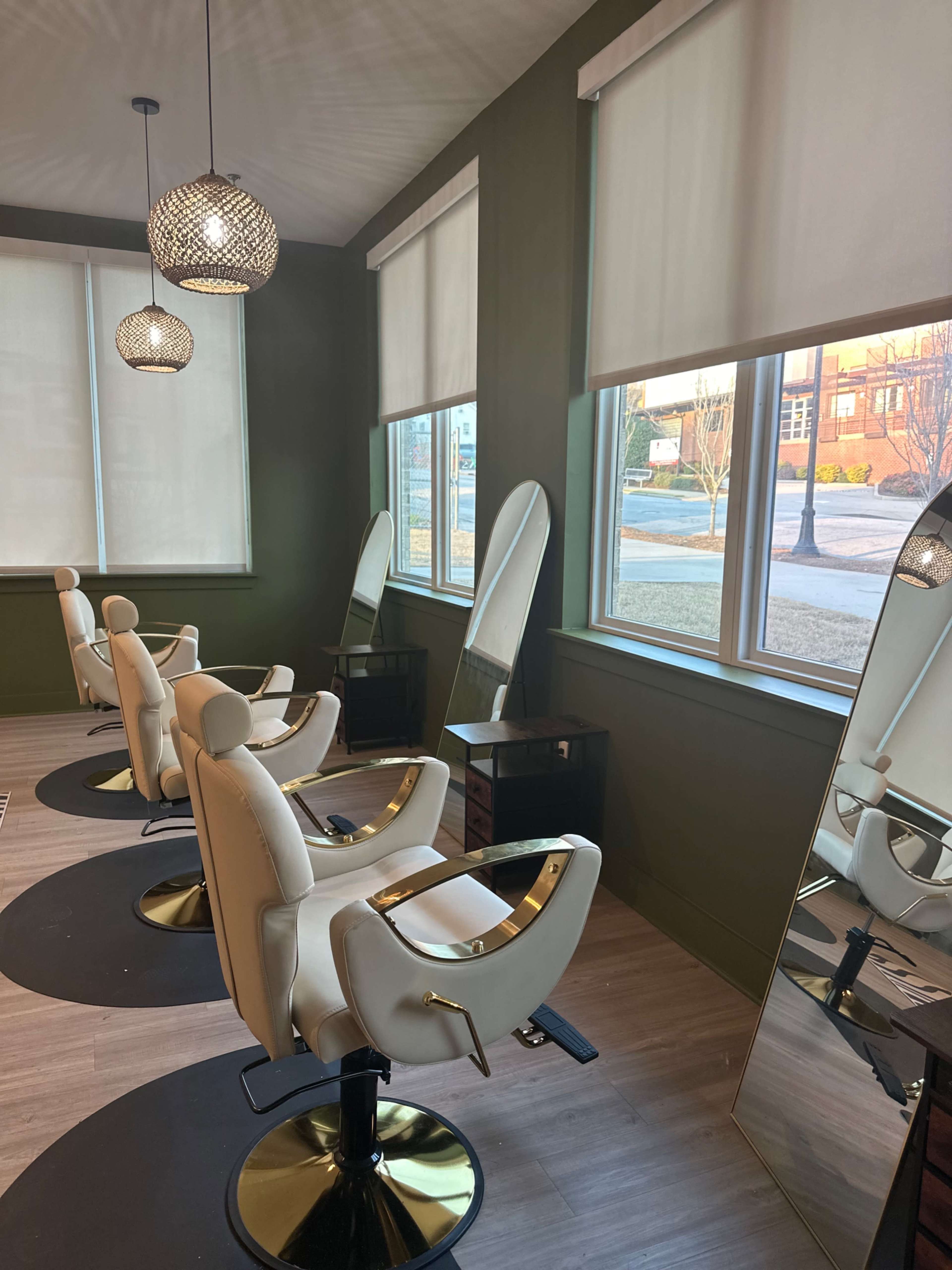 The image shows a modern hair salon interior with three styling chairs facing large windows, illuminated by pendant lighting.