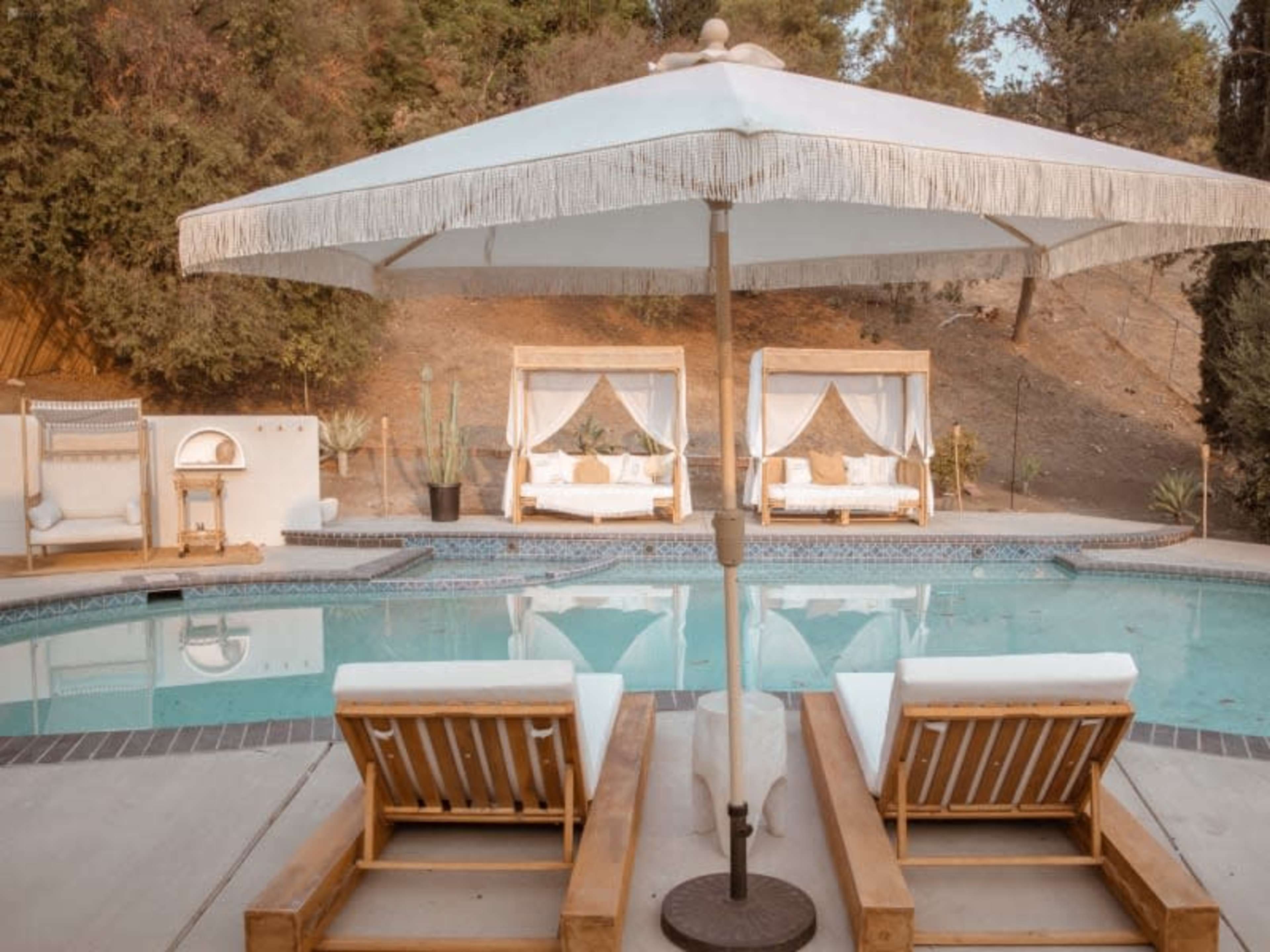A pool area featuring two lounge chairs, a large umbrella, and cabana-style seating surrounded by greenery.