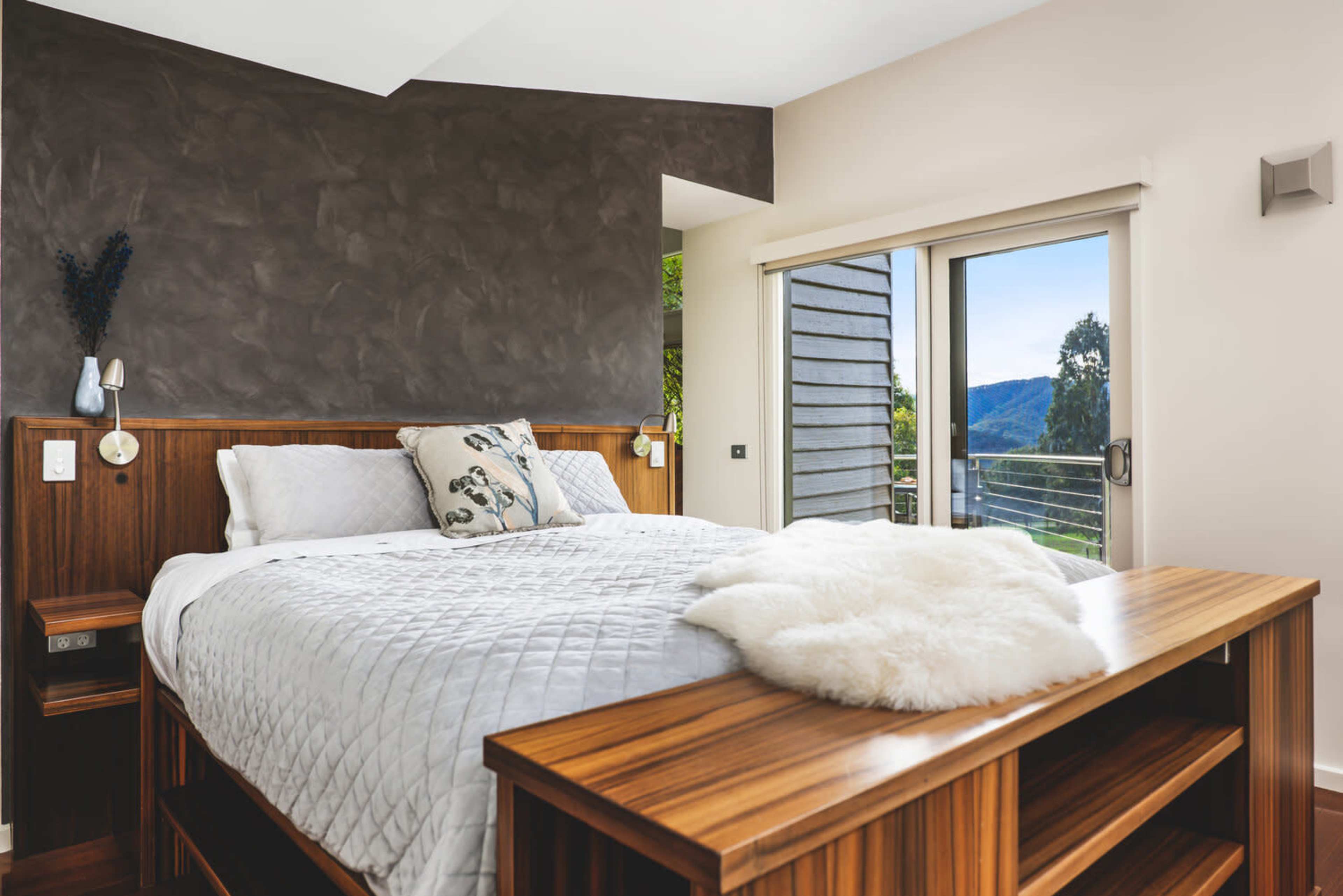 A modern bedroom featuring a large bed with a decorative pillow, a wooden bedside table, and glass doors opening to a view of a mountainous landscape.