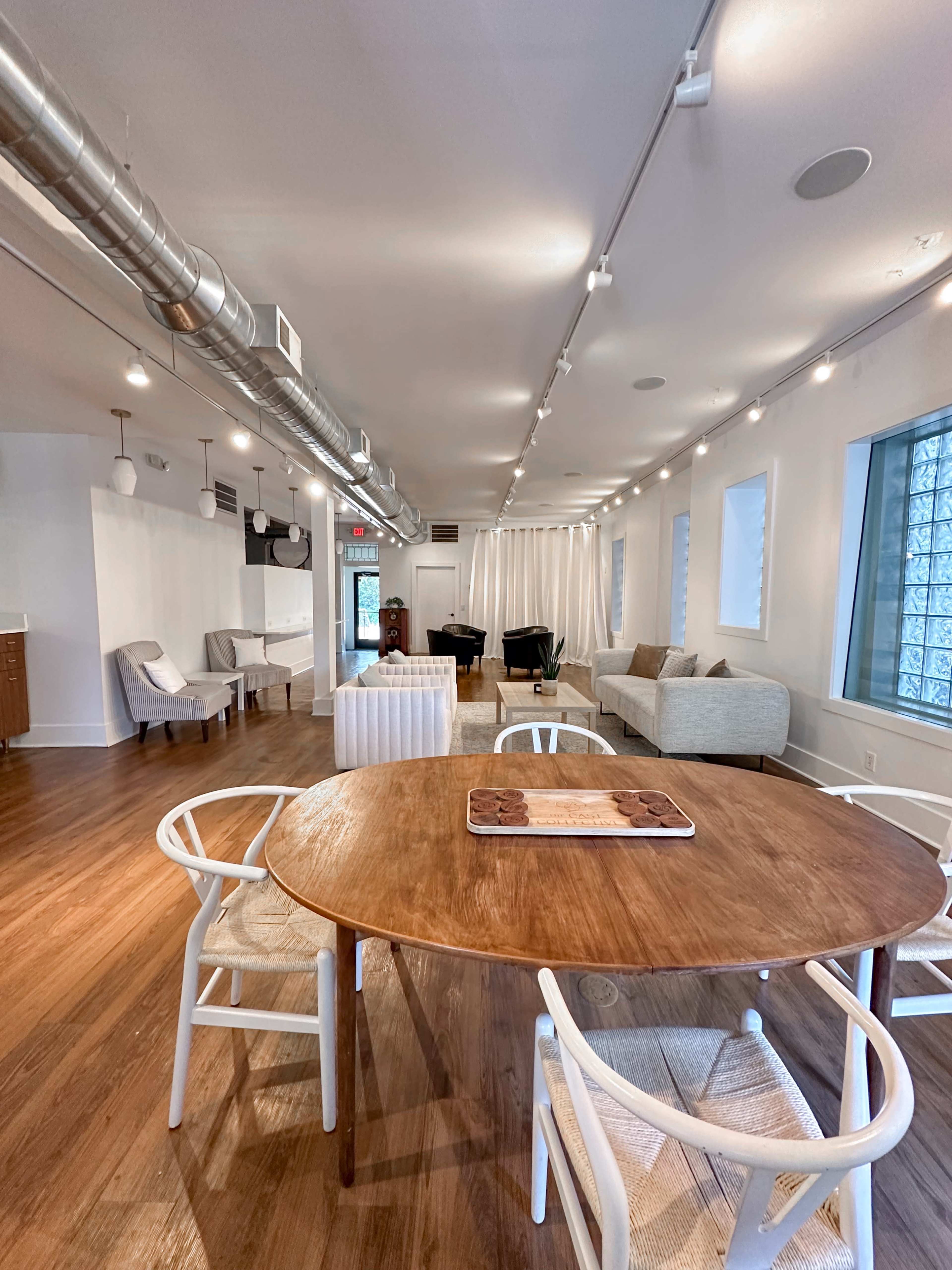 The image shows a modern, airy interior space with a round wooden dining table surrounded by white chairs, along with a lounge area featuring a gray couch and additional seating.