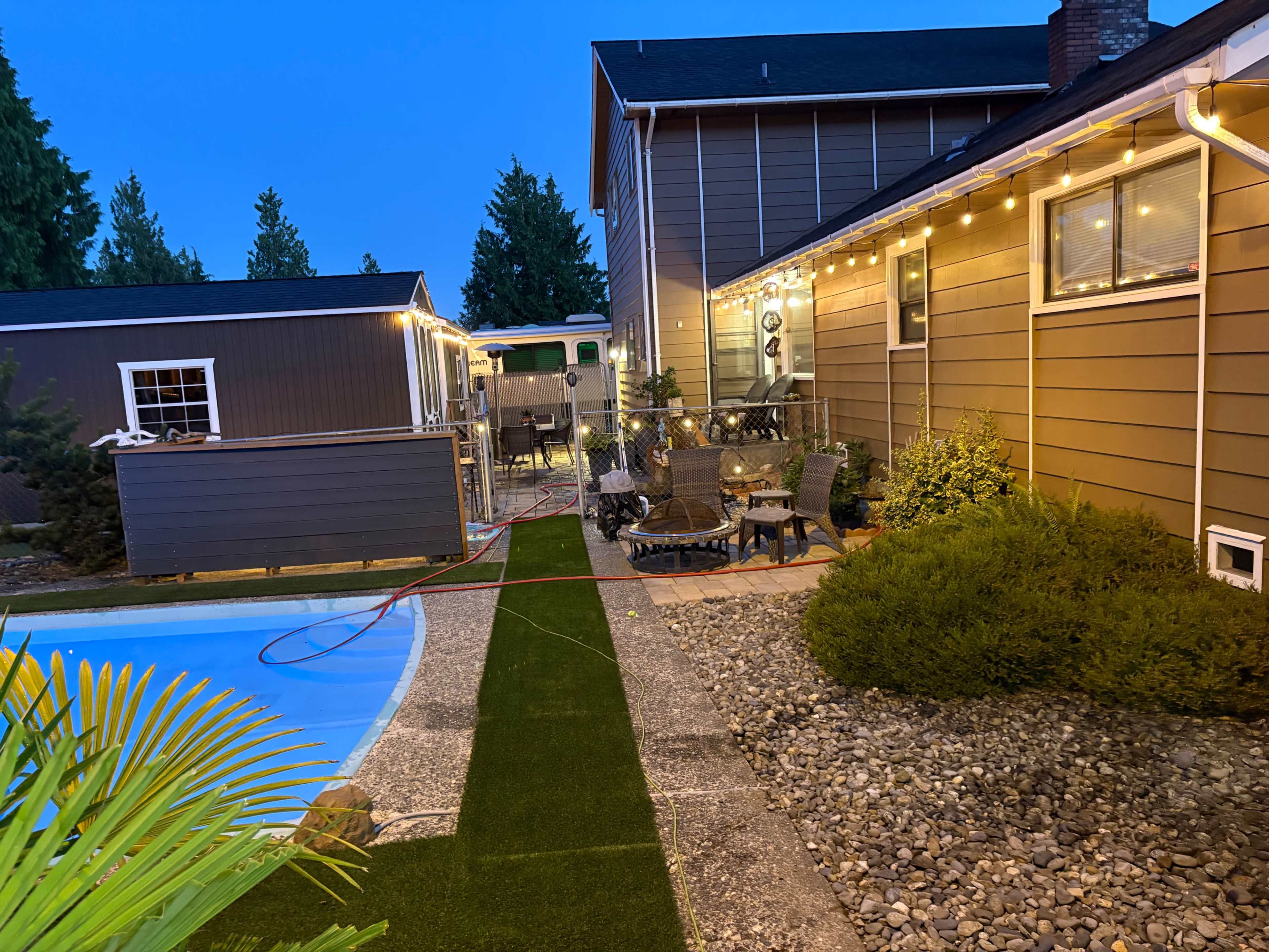 The image shows a backyard with a pool, patio seating area, and decorative string lights on the house, taken during twilight.