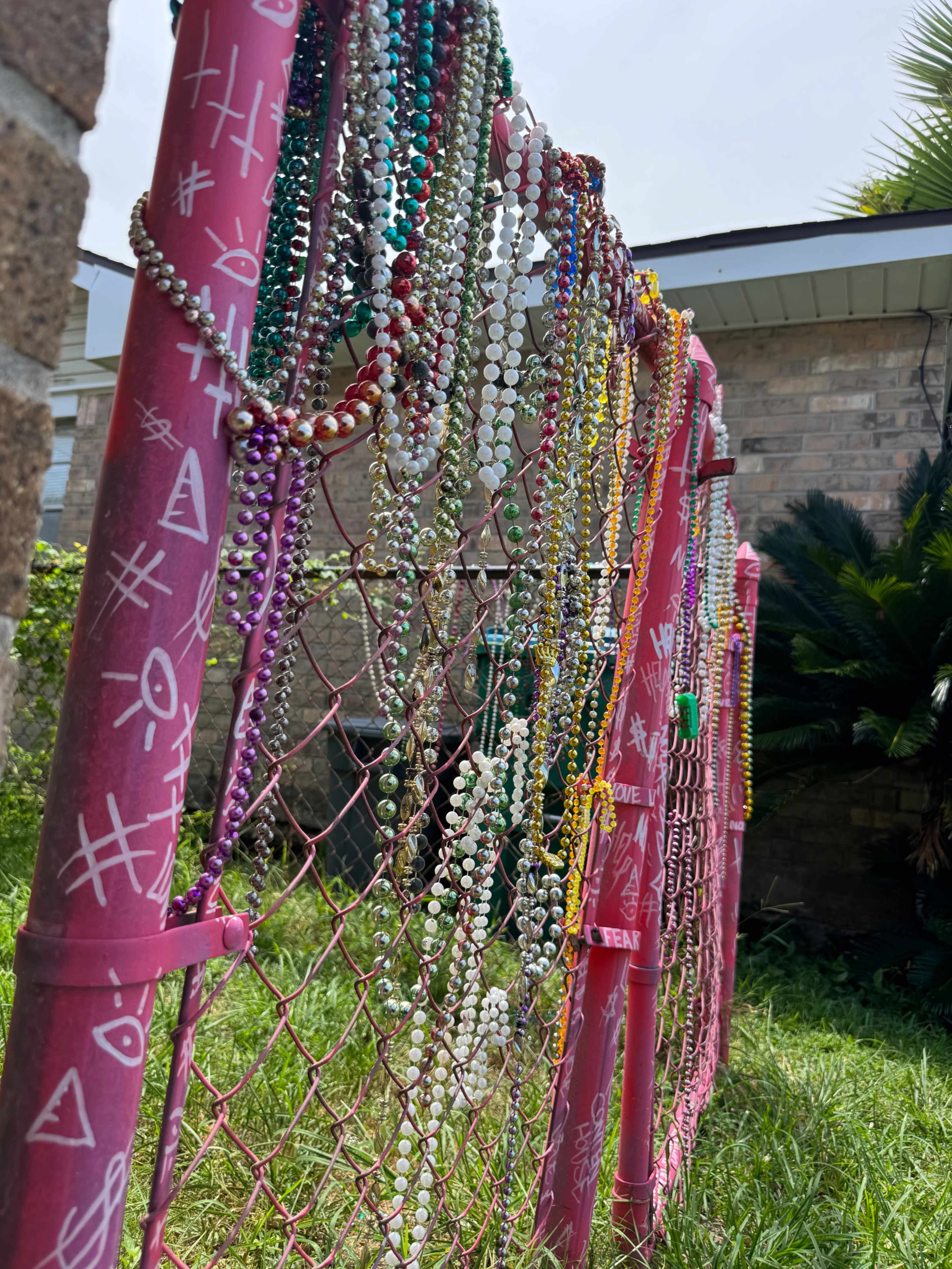 A pink fence is decorated with various strands of colorful beads hanging from its mesh.